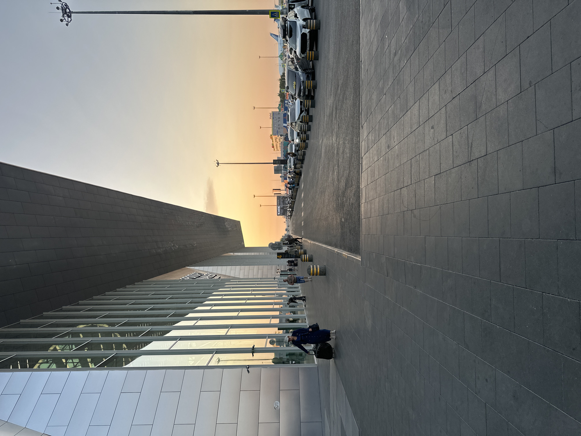 An exterior shot captures the modern architecture of an airport terminal at dusk. The building features a sleek, metallic facade with large, vertical windows reflecting the warm hues of the sunset. A woman in a dark blue dress and a white cap walks along the tiled pedestrian walkway, pulling a black rolling suitcase. She is accompanied by a black tote bag. Other individuals are visible in the background, some walking and others appearing to be employees. Across the street, a line of cars is parked, indicating a pickup or drop-off area. In the distance, airport lights illuminate the tarmac, and an airplane is partially visible. The sky is a gradient of soft orange and pale blue, characteristic of a sunset.