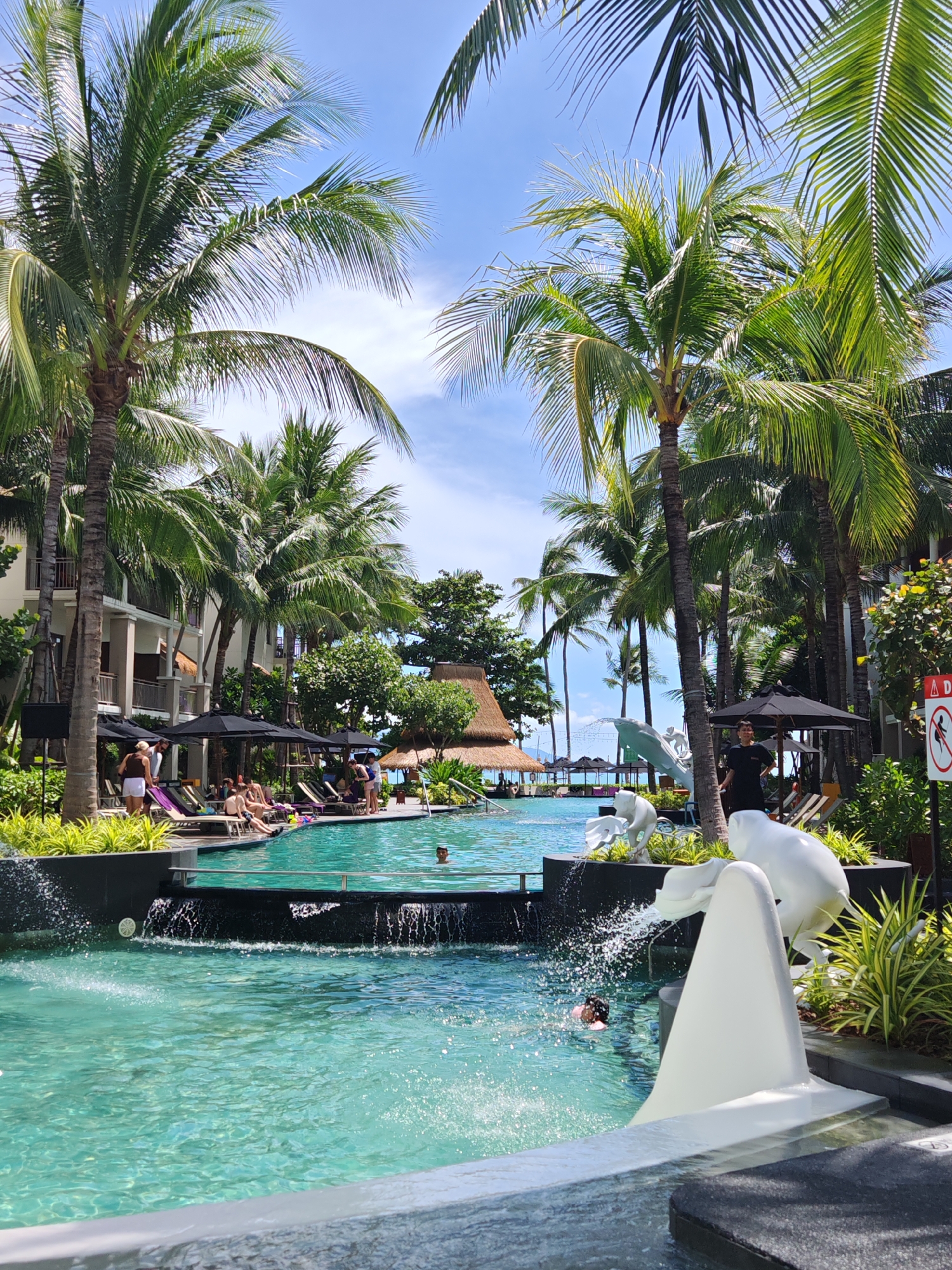 The image depicts a resort swimming pool area in Baan Bo Phut, Thailand. The pool is filled with clear turquoise water, with a cascading waterfall feature at the edge of a raised section. Several white abstract sculptures are placed around the pool, including a dolphin-like figure that appears to be spitting water. Palm trees line the pool and the surrounding area, casting shadows on the water and ground. In the background, there are multi-story buildings with balconies and thatched-roof structures. Several people are visible: some are lounging on poolside chairs under black umbrellas, while others are in the pool. One person is swimming, and another appears to be emerging from the water. A man in a dark t-shirt stands near the sculptures. Lush green vegetation is interspersed throughout the scene. The sky is clear blue with some scattered clouds.