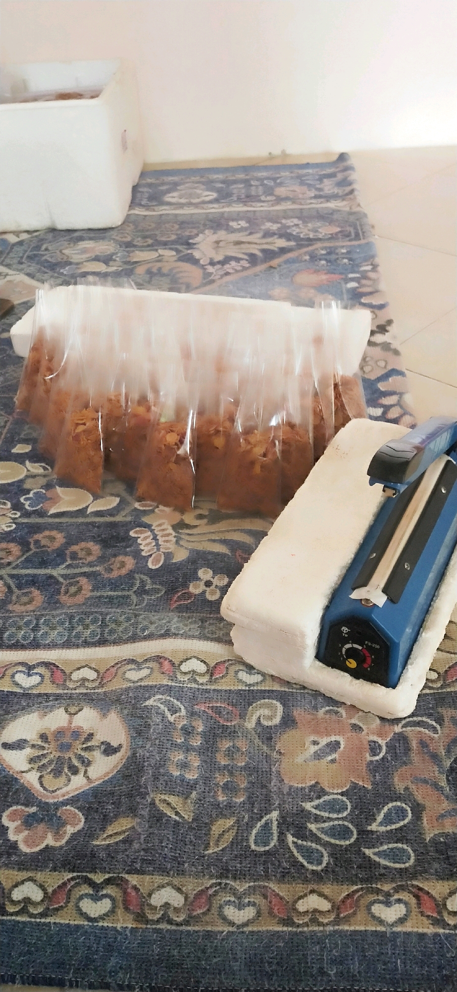 A blue and black impulse sealer sits on a white foam pad, positioned on a patterned rug. To the left of the sealer, a row of food items, possibly baked goods, are individually sealed in clear plastic bags. The bags are arranged in a line and appear to be resting on a white surface, possibly a cooler or container. The rug displays a traditional, intricate floral pattern in shades of blue, tan, and red. The background is a plain, light-colored wall. The scene suggests food preparation or packaging in progress.