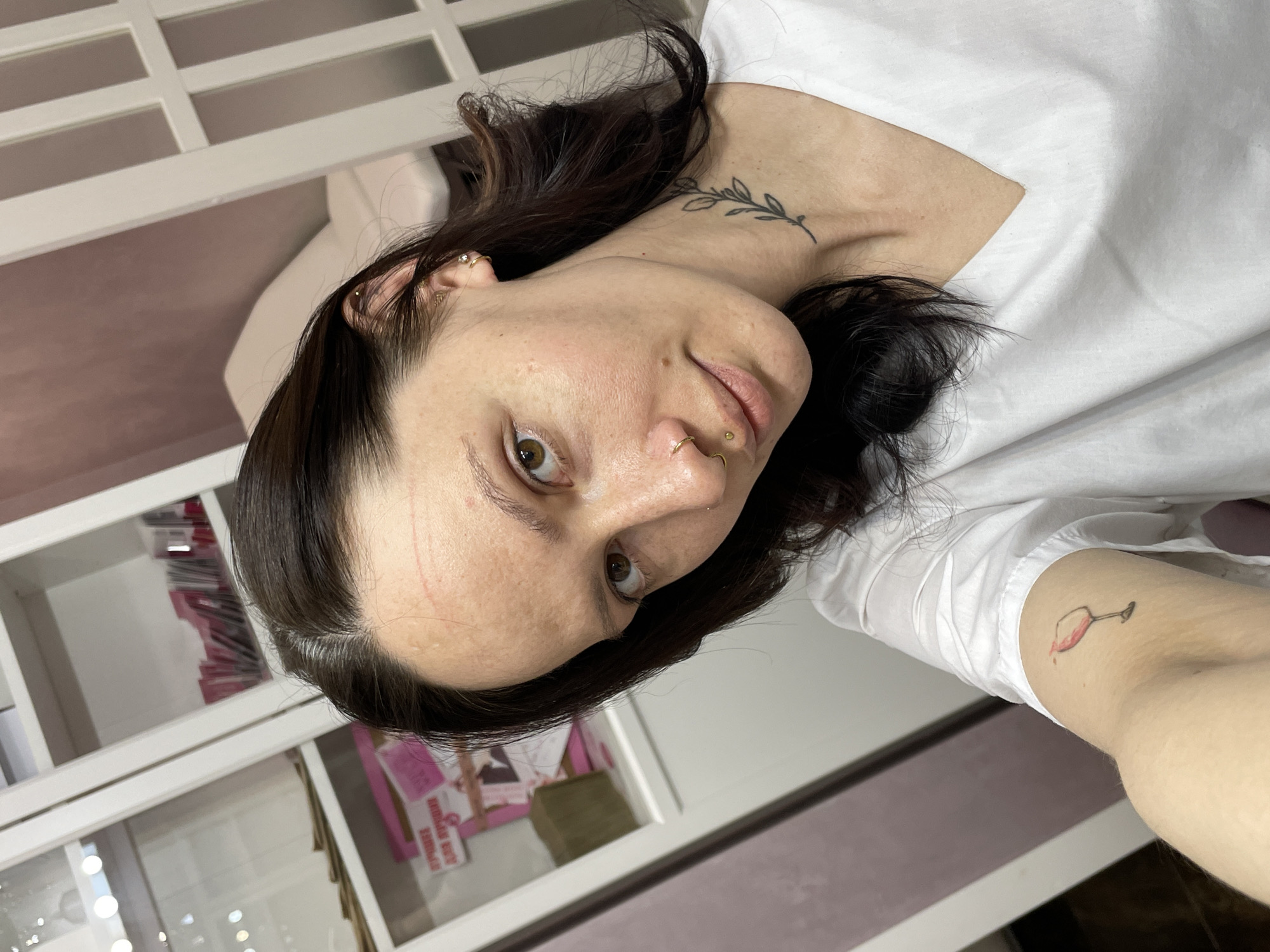 A woman with dark hair is in a selfie-style photo. She has a tattoo of a sprig of leaves on her neck and a tattoo of a wine glass on her arm. She has multiple piercings in her ear and nose. There appear to be scratches on her forehead. She is wearing a white shirt. The background includes white cabinets with contents inside. There is no indication this image was taken in Izhevsk, Russia.