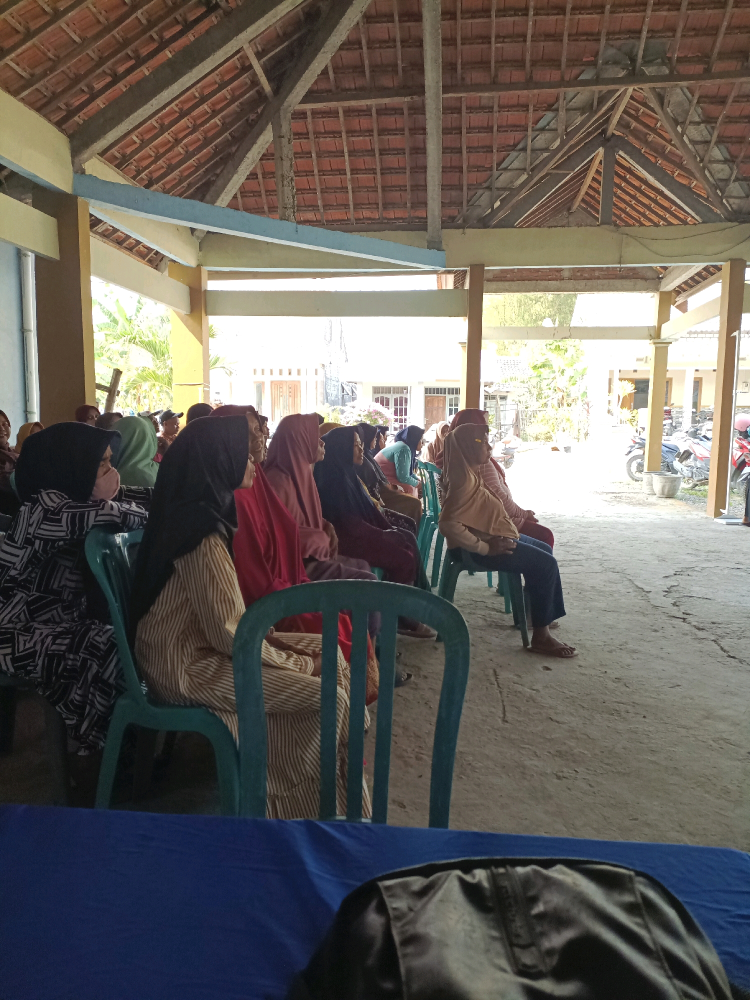 In Rancang Kencono, Indonesia, a group of people, mostly women wearing hijabs, are seated on plastic chairs under a covered outdoor area. The structure has a tiled roof supported by wooden beams and concrete pillars. Some individuals are facing forward, seemingly attending an event, while others are looking to the side. The ground is paved with concrete. In the background, there are buildings and motorcycles parked outside. A blue cloth covers a table in the foreground, and a black bag is partially visible on it. The overall scene suggests a community gathering or meeting.