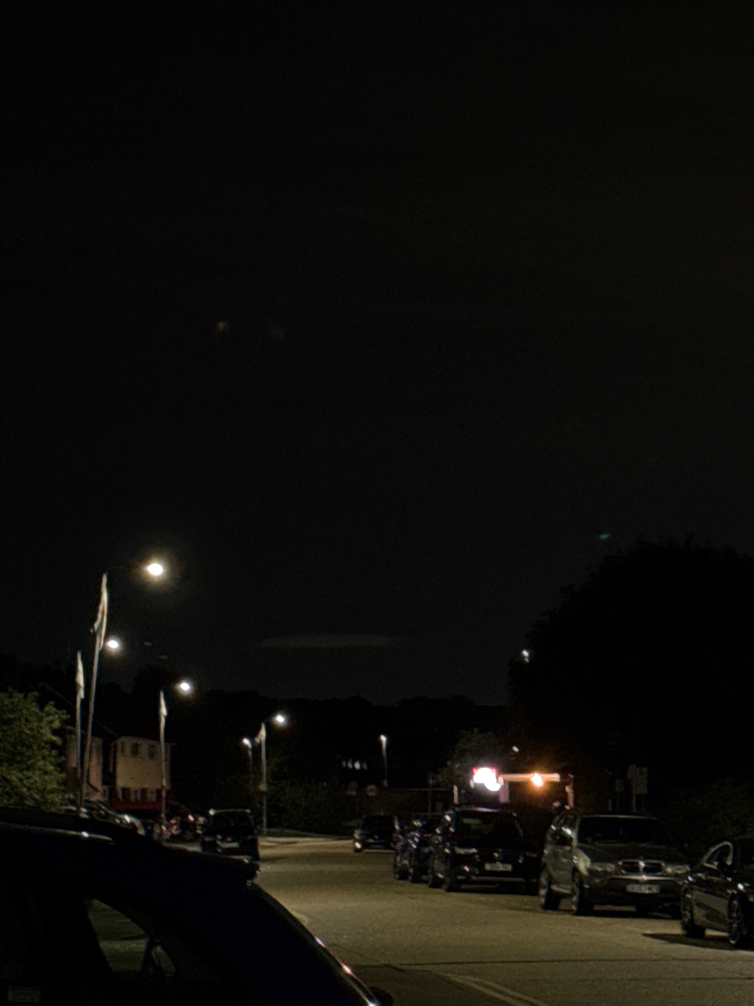 The image is a nighttime scene on a street in Basildon, United Kingdom. The sky is dark, with a bright light that could be the moon or a street lamp visible in the upper left quadrant. Several street lamps are also visible along the road, illuminating the scene.
On the left side of the image, a dark-colored car is parked in the foreground, partially obscuring the view of the street. Beyond this car, there are some buildings and trees. The buildings appear to be houses or shops, with windows that are either dark or illuminated.
The street itself is paved and appears to be a residential or commercial area. Several other cars are parked along the street, including a silver SUV, a dark sedan, and a few other vehicles further down the road. Some of these cars have their headlights on.
In the background, there are more trees and buildings, and the overall impression is of a quiet, urban environment at night. There are no people visible in the image. The lighting is low, creating shadows and a somewhat grainy quality to the image.