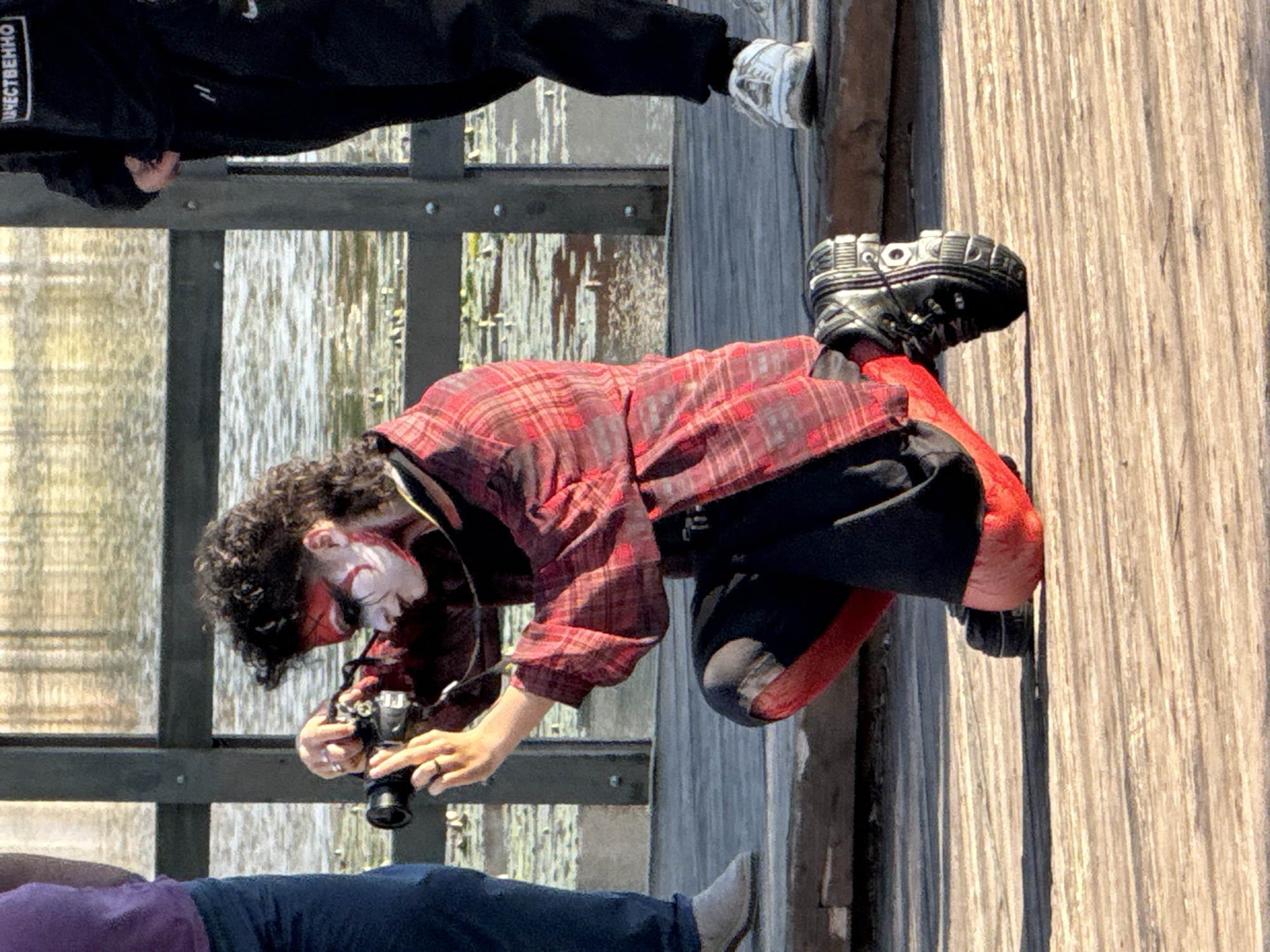 A person with dark, curly hair and white and red face paint is kneeling on a wooden boardwalk, holding a camera to their face with both hands. They are wearing a red and black plaid shirt, black pants with red accents on the legs, and black boots with white laces. The person appears to be taking a photograph. In the background, there is a body of water with reflections of trees and a railing. To the left, a person in a purple shirt and blue jeans stands. To the right, a person in black pants and white sneakers stands with their back partially to the camera. The scene appears to be outdoors, possibly near a lake or river. The location is specified as Votkinsk, Russia.