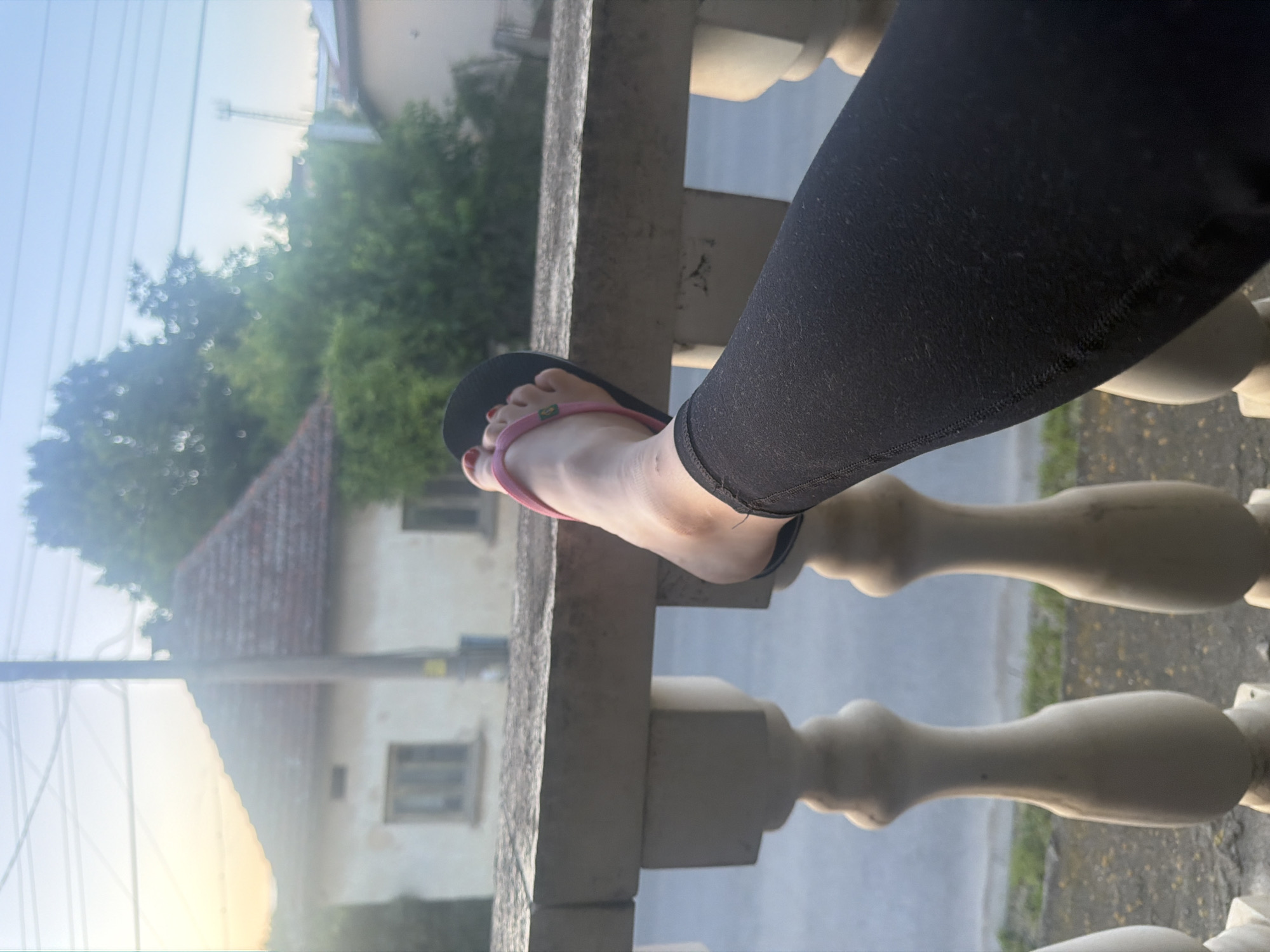 A slightly tilted image captures a person's left leg and foot resting on a stone balcony railing. The foot is bare, showing red painted toenails, and is partially clad in a black flip-flop with a bright pink strap. A small, indistinct green logo is visible on the pink strap. The person is wearing black leggings, which appear to be made of a textured fabric, with visible stitching at the ankle cuff.
The setting is outdoors, suggesting a balcony or terrace overlooking a residential area. The background is softly blurred, but details such as green trees and a house with a distinctive reddish-brown tiled roof and light-colored walls are discernible. The sky above is clear and light blue, indicating fair weather, likely late afternoon or early evening due to the soft, bright illumination. Power lines are visible stretching across the upper left portion of the sky. The railing itself is made of light-colored stone or concrete, featuring decorative balusters. The scene conveys a sense of calm and relaxation.
