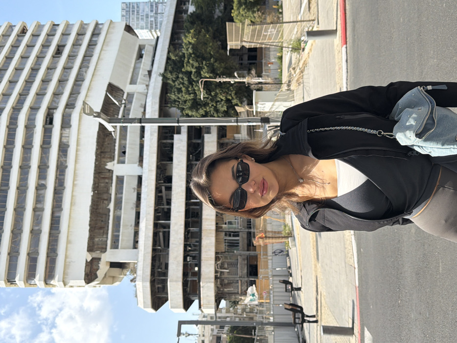 The image displays a young adult female standing on a paved street or sidewalk in Tel-Aviv, Israel, with the prominent, dilapidated former Dizengoff Square Hotel (also known as the Astoria Hotel or Hotel Center) in the background. The woman is centered, facing slightly left of the camera, and looking forward. She has brown hair and wears dark, rectangular sunglasses, a black zip-up jacket over a dark grey wrap-style top and a white undershirt, and dark grey fitted pants. She accessorizes with a silver-colored necklace with a small light-colored pendant and carries a light blue denim-textured mini backpack adorned with silver studs and a chain strap on her left side.
Behind the subject, the multi-story Dizengoff Square Hotel exhibits a light-colored, weathered facade with significant damage and exposed internal structures across multiple floors, particularly the lower sections. Its architecture features a tiered, angular design with numerous empty or broken window openings. To the left of the woman, beyond the street, a sidewalk contains brown benches and temporary construction barriers. A tall black street lamp post is situated directly behind the woman, with another visible further left. Green trees are positioned to the sides of the large background building. The sky is clear blue with minimal white clouds, indicating daylight conditions.