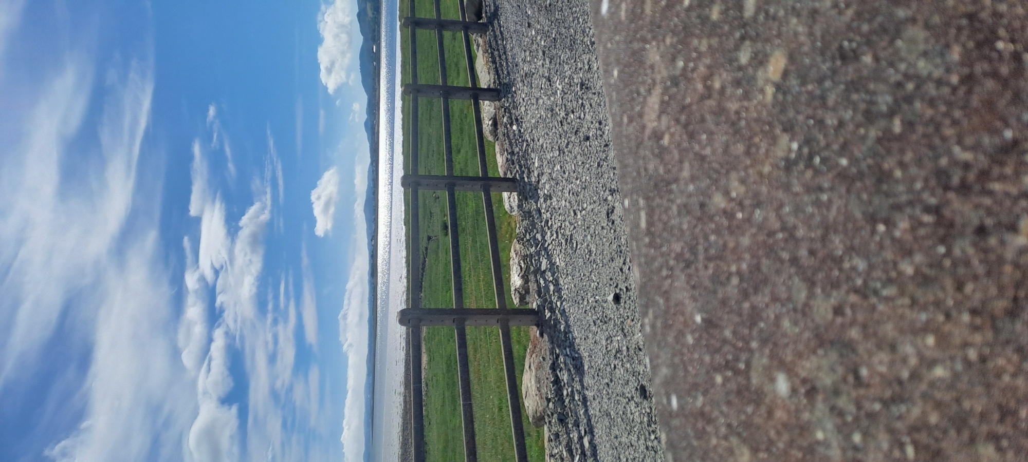 This image depicts a scenic view from Pier Road in Ireland, likely taken on a sunny day with scattered clouds. The foreground is dominated by a rough, gravelly surface, possibly a path or embankment, with a rustic wooden fence bordering it. Beyond the fence, a vibrant green grassy field stretches out, meeting a calm body of water. The water's surface is highly reflective, catching the bright sunlight and creating a shimmering effect. In the distance, a hazy mountain range or hills form the horizon line, partially obscured by the bright sky. The sky itself is a brilliant blue, adorned with wispy white clouds that are characteristic of fair weather. The composition is oriented vertically, suggesting the photo was taken with a phone held in portrait mode, and then rotated. There is no visible text in the image. The overall atmosphere is peaceful and serene, evoking a sense of tranquility associated with a coastal or lakeside landscape. The time of day appears to be daytime, likely late morning or early afternoon, given the brightness of the sun. The location cues, such as the fence, grassy field, water, and distant hills, are consistent with a rural or semi-rural setting in Ireland, as suggested by the provided location context. There are no people or specific objects beyond the natural elements and the fence.