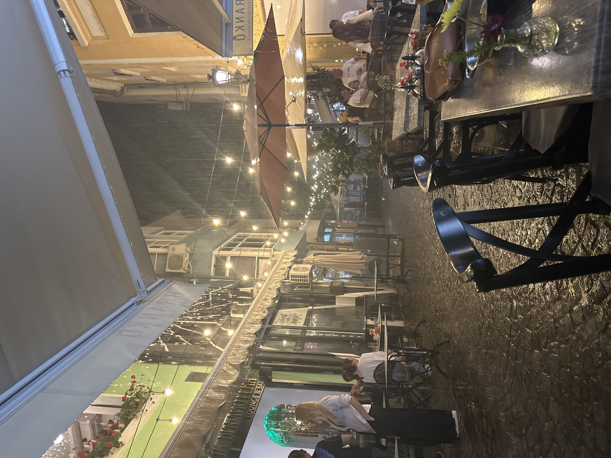 This image captures a rainy night scene on a pedestrian street in Plovdiv, Bulgaria. Heavy rain is actively falling, characterized by visible streaks through the air and numerous reflective wet surfaces, including the cobblestone ground, awnings, and umbrellas.
The street is illuminated by multiple strings of warm-toned bulb lights strung overhead, supplemented by interior lighting from businesses and a decorative street lamp on the right side.
On the left, a business identified as "БИБЛИОТЕКА LOUNGE & BAR" features a green-painted facade, large windows, and a light-colored awning from which water drips. A window box or balcony above displays red flowers. A blonde woman in a white shirt and black skirt stands near the entrance, facing away from the camera, interacting with a man. Two other women are seated at a table directly outside this establishment.
Further down the street and to the right, a cream-colored building features an awning with the reversed Cyrillic text "ОКНАРН." An outdoor air conditioning unit is mounted on a building wall near the center.
Approximately 10-15 individuals are present. Several patrons are seated at dark-colored outdoor tables and chairs, primarily under large, open, tan-colored patio umbrellas that are visibly shedding rainwater. One group includes a man in a white t-shirt, a woman, and a small child.
In the immediate foreground, a wet wooden table displays small glass vases containing flowers, a salt and pepper shaker set, and a brown leather handbag. The entire street surface is composed of dark, wet cobblestones, reflecting the overhead lights intensely.