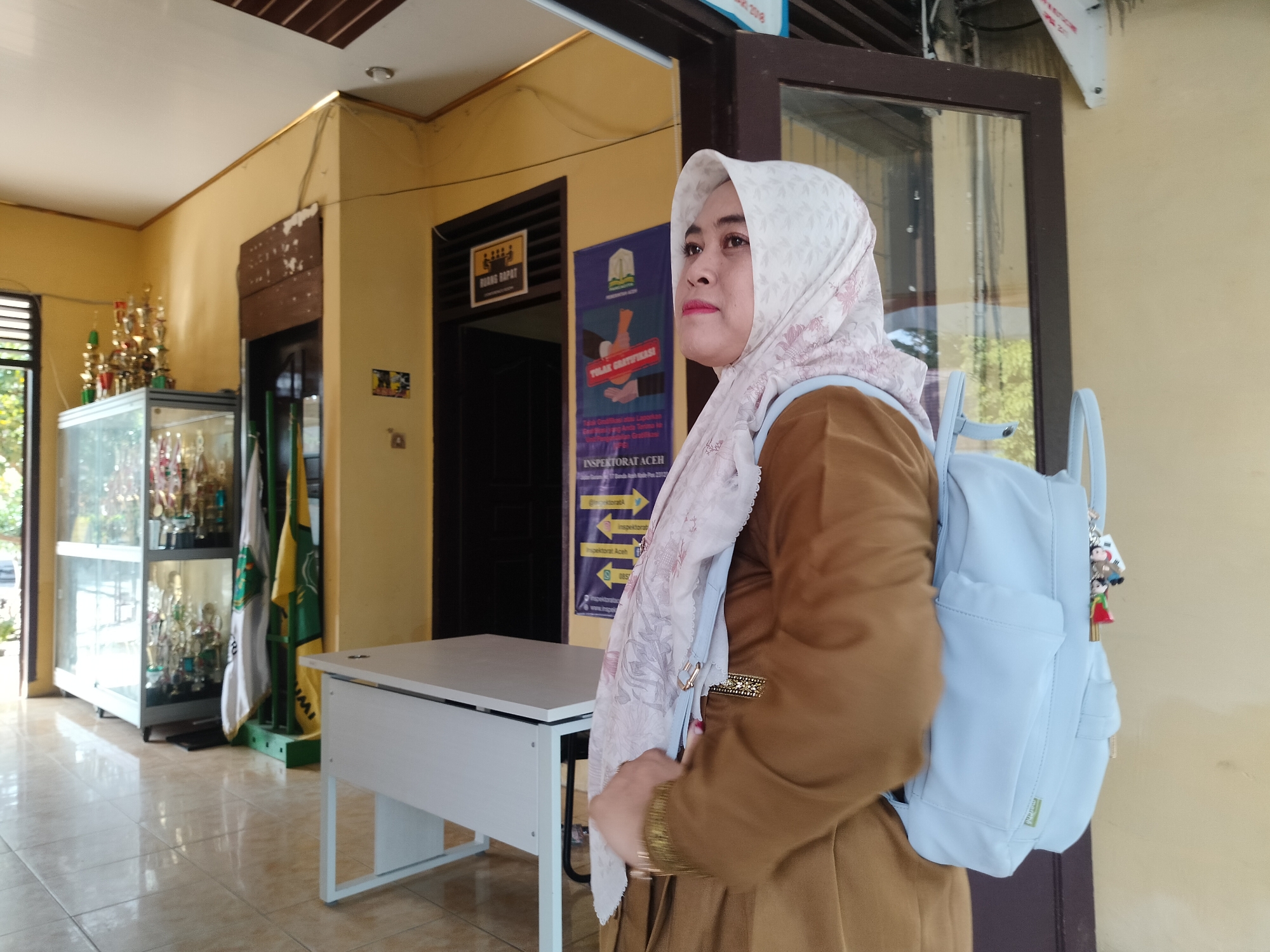 A medium shot captures a woman wearing a brown long-sleeved tunic and a white patterned hijab. She has a light blue backpack with two cartoon charms on the strap. The woman is standing in front of a doorway, looking to her left. Behind her is a yellow wall, a closed door with a sign that reads "RUANG RAPAT" (Meeting Room) and another sign that is partially visible, and a poster. To her left, there is a glass display case filled with trophies, flags, and a white desk. The floor is tiled and reflective. The environment appears to be an office or administrative building, possibly within Lawebekung, Indonesia, given the text on the signs. The lighting suggests an indoor setting with natural light coming from the open doorway.