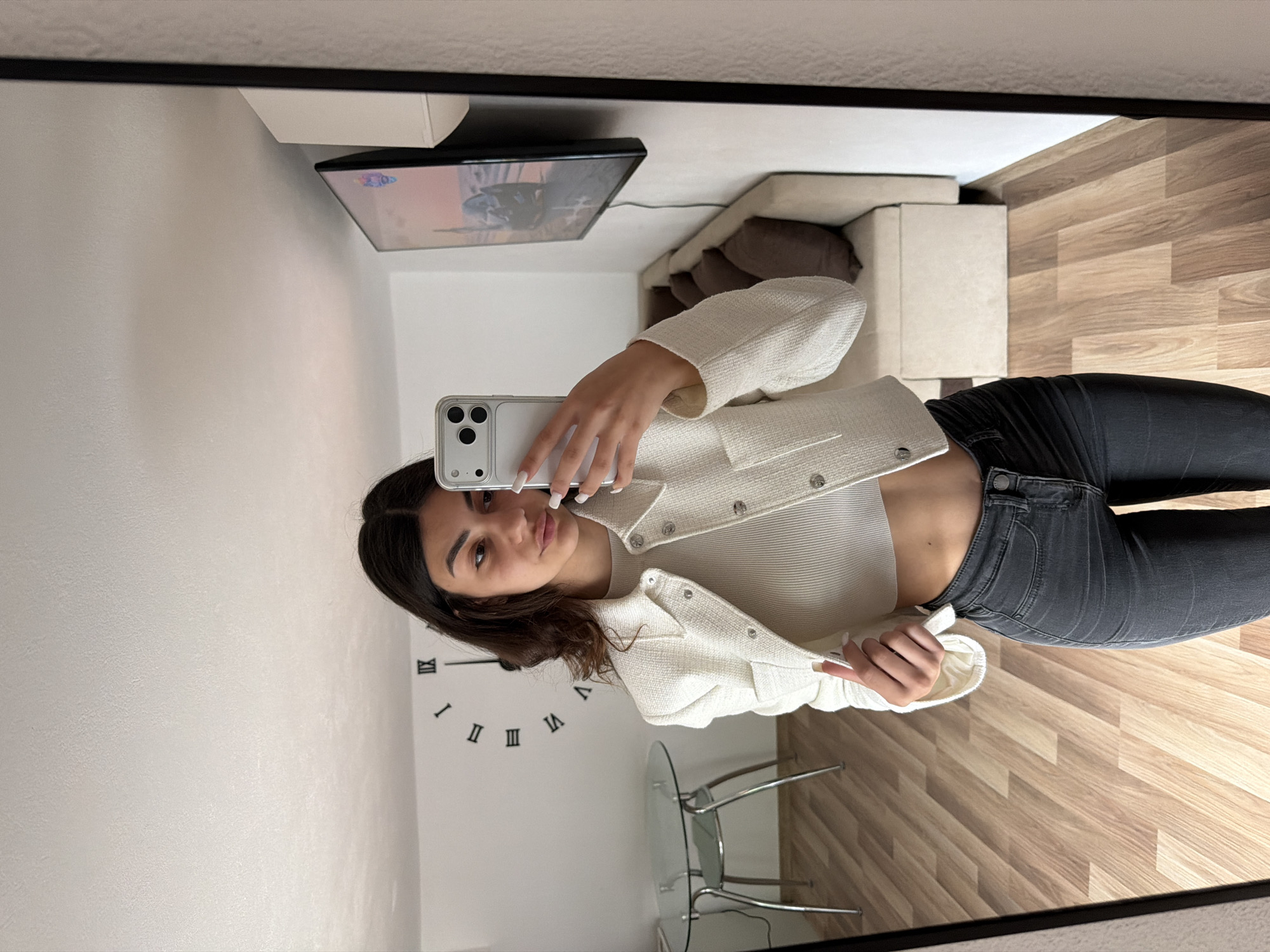 A young woman takes a mirror selfie in what appears to be an apartment setting. The background features light-colored walls, wooden flooring, and furniture including a sofa. A wall clock is visible behind the subject. There's also a glass table with metal legs. The scene isn't identifiable within Gabrovo, Bulgaria without additional contextual clues.