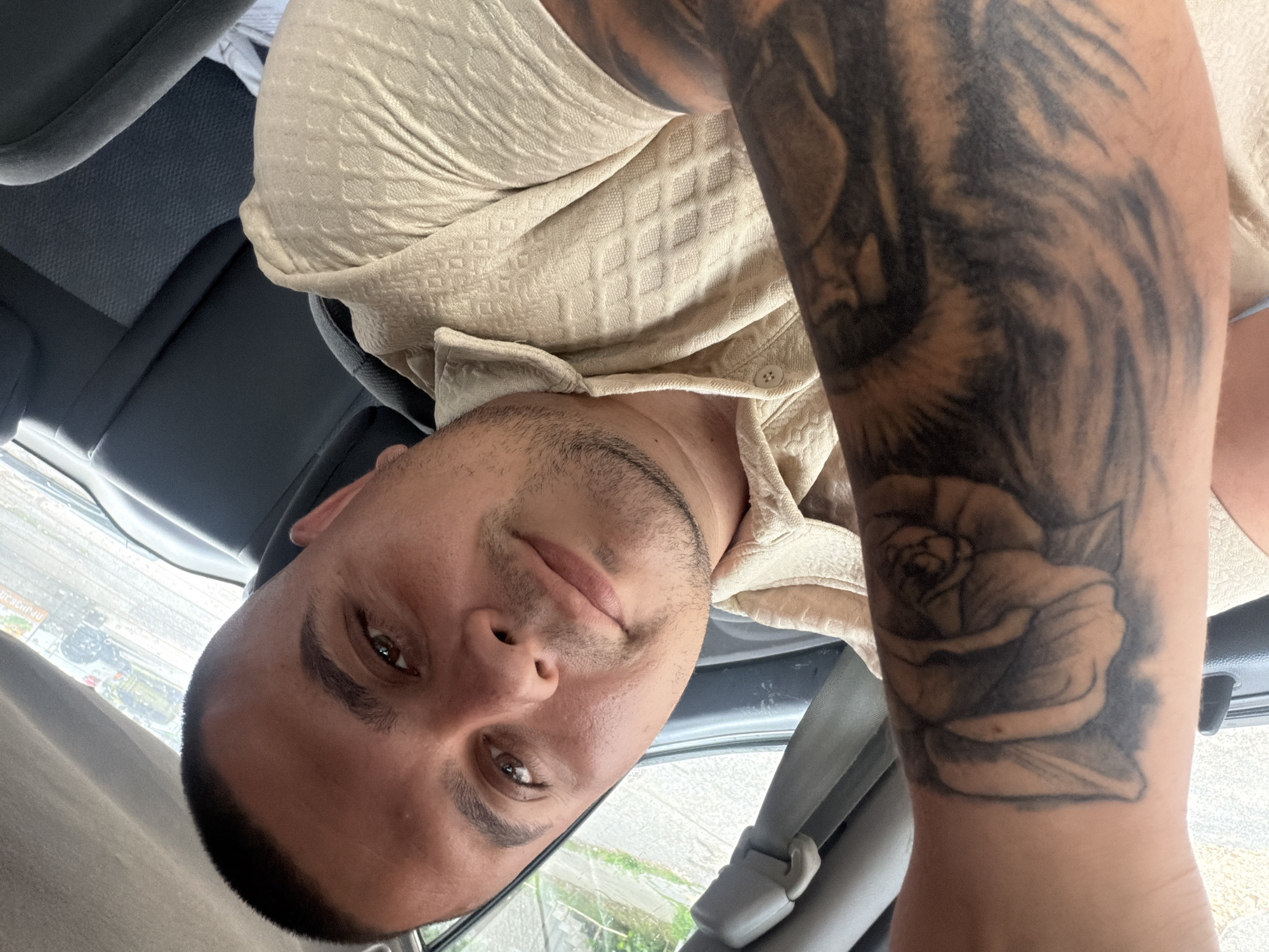 A male individual, estimated to be in his late 20s to early 30s, is captured in a self-portrait within the interior of a vehicle in Shumen, Bulgaria. He has short, dark, faded hair and dark, thick eyebrows, with a light stubble on his chin and jawline. His brown eyes are directed towards the viewer, and he exhibits a subtle, relaxed smile. He is wearing a light beige, short-sleeved collared shirt with a textured pattern resembling small diamonds or squares.
His left forearm, prominently featured in the foreground, is extensively tattooed. The upper portion of the forearm displays a large rose or floral design with intricate shading, while the lower part of the forearm near the wrist features a pattern consistent with tiger stripes.
The vehicle's interior is visible, featuring dark grey upholstery and plastic trim. A seatbelt mechanism is discernible on the upper left. Through the windows, an outdoor urban or semi-urban environment is visible. The right rear window shows a street scene with faint reflections of trees and buildings, along with visible raindrops on the glass, suggesting recent precipitation. A partially visible, faded orange rectangular sign with Cyrillic script is discernible in the background, consistent with signage found in the city of Shumen, Bulgaria. The overall lighting suggests daytime conditions, possibly overcast.