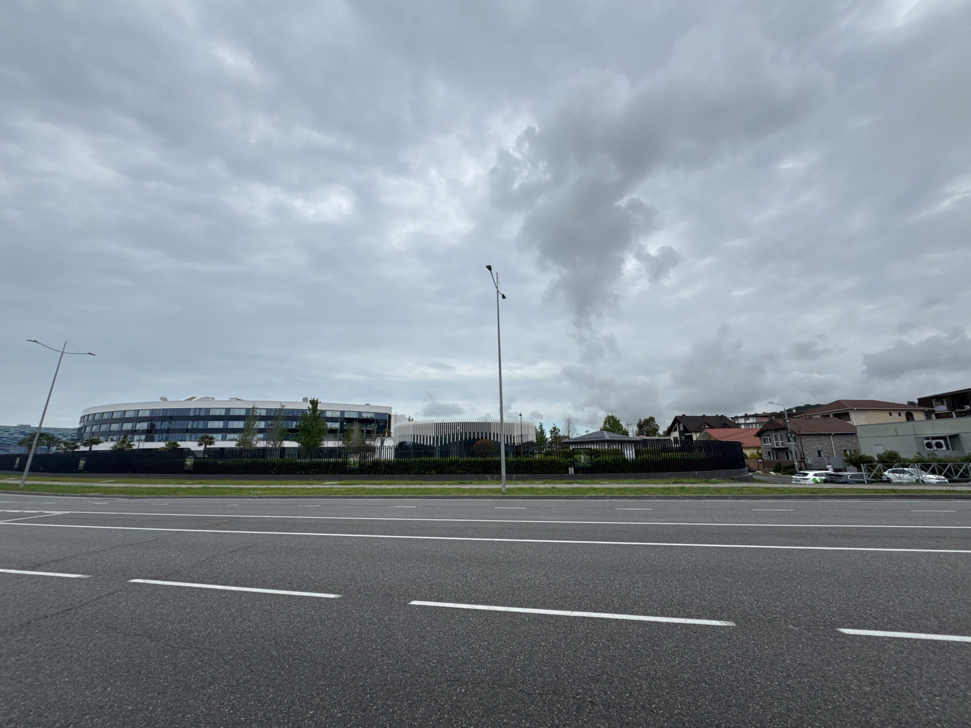 The image depicts a street scene in Sirius, Russia, under a cloudy sky. An asphalt road with white lane markings dominates the foreground. Behind a low green hedge and black fence are two modern buildings. The building on the left is curved and has multiple stories with reflective windows. The building on the right is also modern and has a white façade with vertical lines. Further back, residential buildings are visible on a slight hill, and several parked cars are discernible. Two streetlights stand between the road and the buildings. No people are visible.