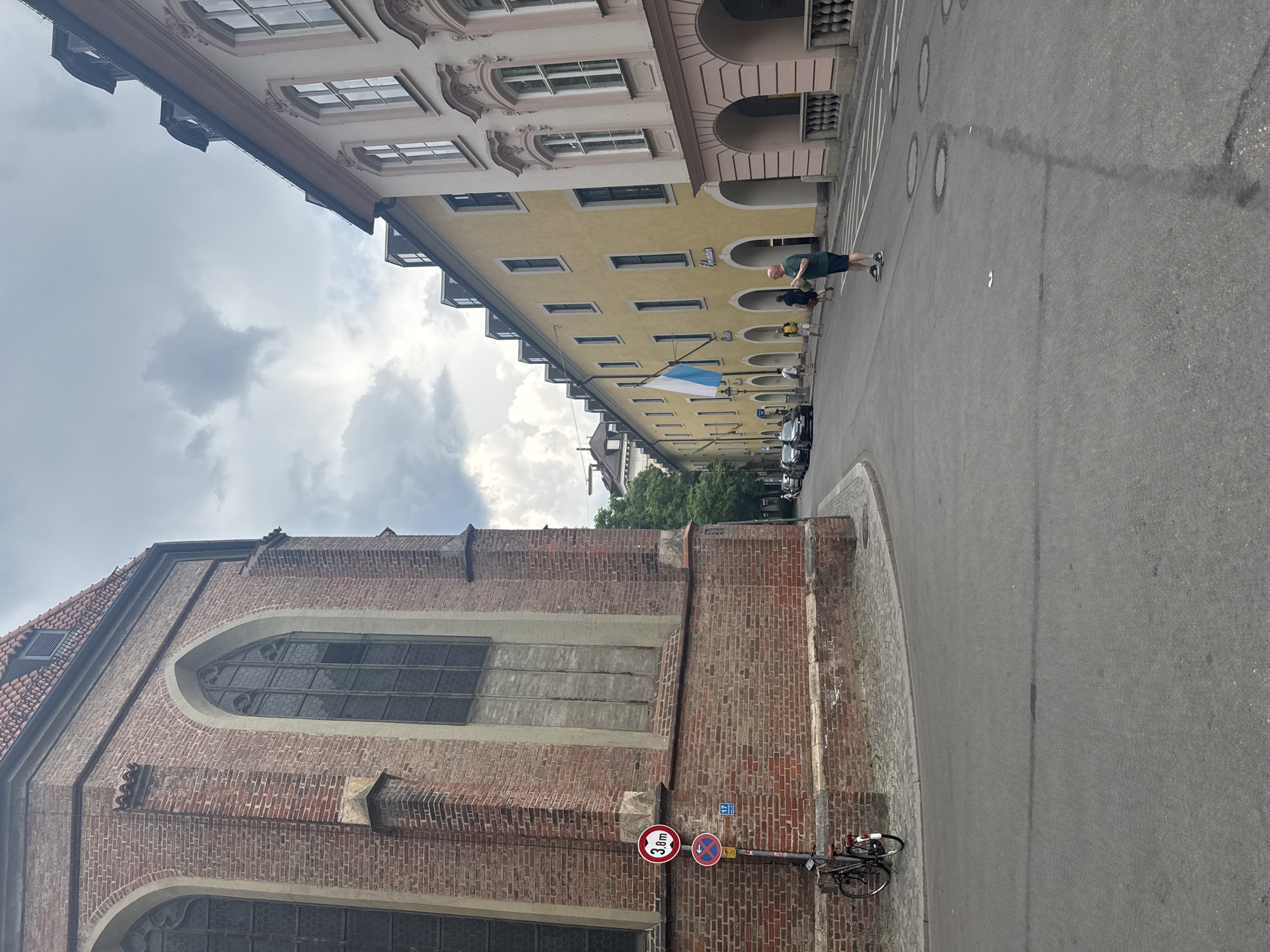 The image captures a street scene in Munich, Germany, with a prominent brick building on the left, identified as a church due to its Gothic windows. The building features a red tile roof and arched windows with dark panes. In front of the church, a bicycle is parked next to a pole with traffic signs, including a circular red sign with "3.8m" and a blue sign with a left arrow and the number "17".
To the right of the church, a street runs along a row of buildings. The closest building is a pale yellow structure with arched ground-floor openings and regularly spaced windows on upper floors. A Bavarian flag, blue and white, is displayed from this building. Further down the street, a lighter-colored building with classical architectural elements is visible. Several people are walking on the sidewalk and in the street. One person, wearing a green shirt and dark shorts, stands in the street looking down. The sky is overcast with clouds. Cars are parked and driving on the street.