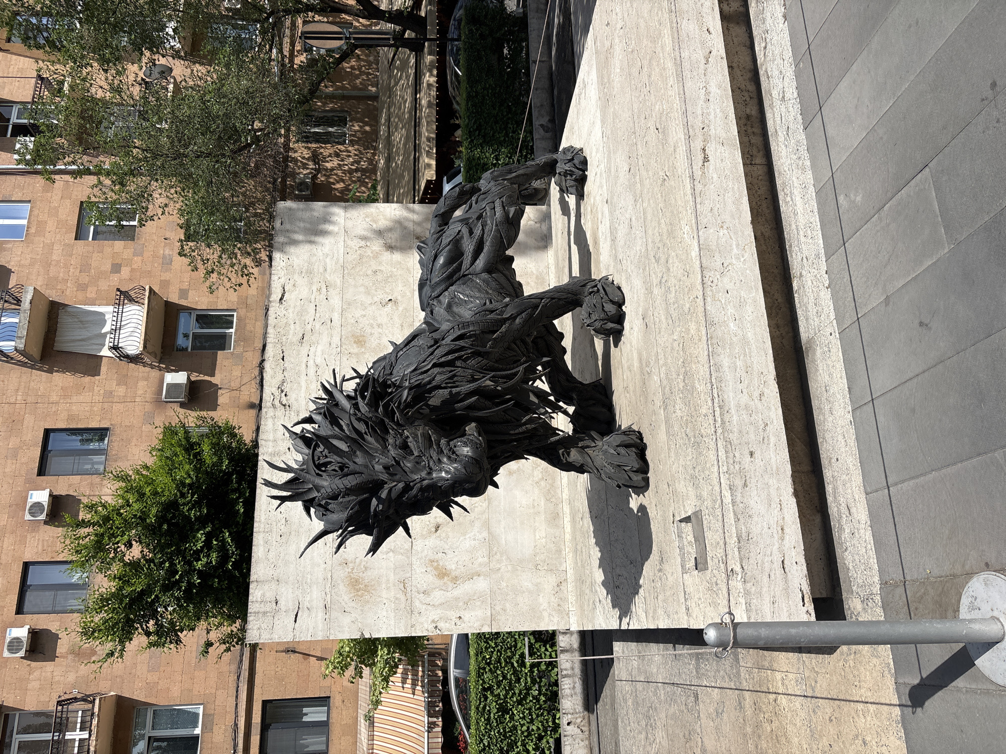 A sculpture of a lion, made from black tires, stands on a stone pedestal in front of a tan building with a textured facade. The lion sculpture is dark and appears to be crafted from cut and assembled tire pieces, giving it a rough, spiky texture, particularly around its mane and tail. Its body is muscular and detailed, with its head lowered and facing slightly to the left.
The pedestal is made of light-colored, textured stone blocks and is situated on a paved area. Behind the sculpture, a light-colored stone wall rises, with a portion of the tan building visible above it. The building has multiple windows, some with black frames and balconies with white railings. Several air conditioning units are mounted on the building's facade. Green trees and bushes are visible in the background, some growing behind the wall and near the building. An awning with orange and white stripes is also visible on the left side of the image, with a car parked behind a hedge. The ground in front of the pedestal is paved with gray square tiles. A metal bollard with a rope attached is positioned to the left of the sculpture.
The scene appears to be outdoors in a city environment, likely a public space. The lighting suggests it is daytime, with shadows cast by the sculpture and other elements. This sculpture is located in Yerevan, Armenia, specifically at the Cascade, a giant stairway in the Kentron district of the city.