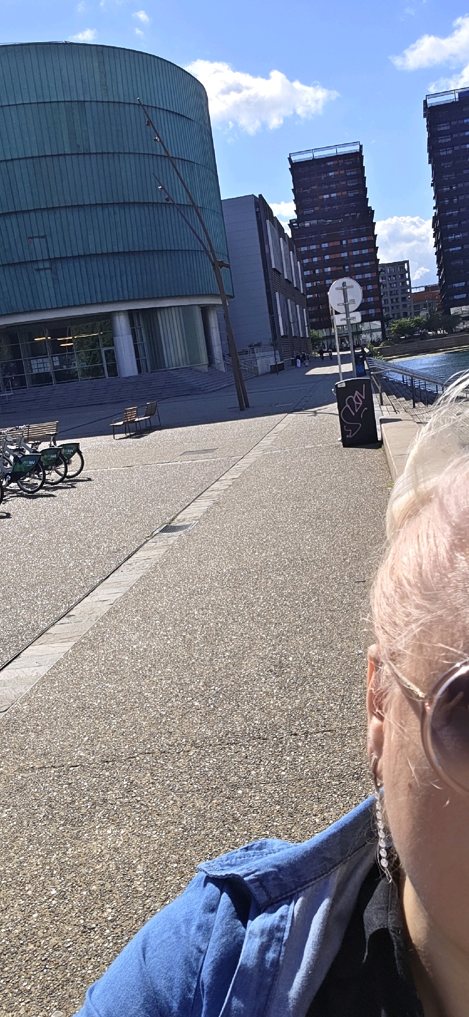 The image captures a sunny day in Strasbourg, France, likely near the European Parliament. In the foreground, the side profile of a person wearing a denim shirt and sunglasses is visible, with their blonde hair illuminated by the sun. Behind them, a textured concrete walkway leads to a modern, cylindrical building with a teal facade, possibly a part of the European Parliament complex. Further in the background, tall, dark, modern apartment buildings are visible, along with a body of water and a railing. Several bicycles are parked to the left of the walkway, suggesting a public space. A trash can with graffiti is also present. The sky is blue with scattered white clouds.