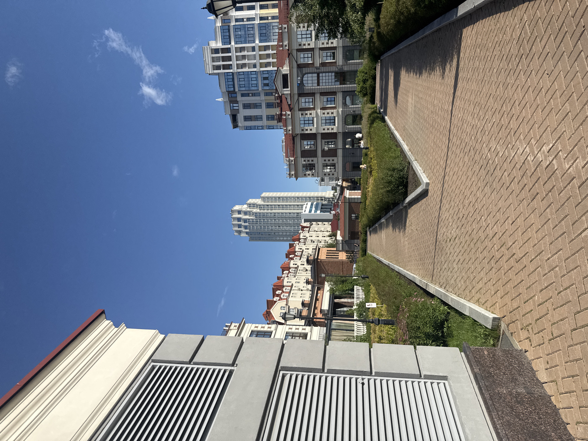 This is an outdoor, eye-level, wide-angle shot of a paved walkway alongside manicured green bushes and a modern building complex under a clear blue sky. The walkway is paved with light brown bricks laid in a herringbone pattern. The buildings are modern, with many windows and balconies, varying in height and architectural style. Some buildings have red tile roofs, while others are contemporary glass and steel structures.
The scene appears to be in a developed urban or suburban area, possibly a residential complex or a business district. There are no people visible in the image. The time of day is likely midday, given the bright sunlight and distinct shadows cast by the buildings and the lamp posts along the walkway. The weather is clear and sunny, with a few wispy white clouds in the sky.
Notable details include the architectural styles of the buildings, ranging from traditional European influences with pitched roofs to sleek, modern high-rises. The landscaping is well-maintained, with neatly trimmed hedges and some flowering plants. A black lamppost is visible along the walkway. There is also a signpost with a small white sign. The foreground shows a close-up of a ventilation grate and a textured concrete structure, suggesting an underground utility access or a decorative architectural element.
Visible text includes "ВТБ" (VTB) written on one of the buildings, which is the name of a Russian bank. The overall mood is serene and orderly, suggesting a well-maintained public or private space. The location context is Yekaterinburg, Russia.