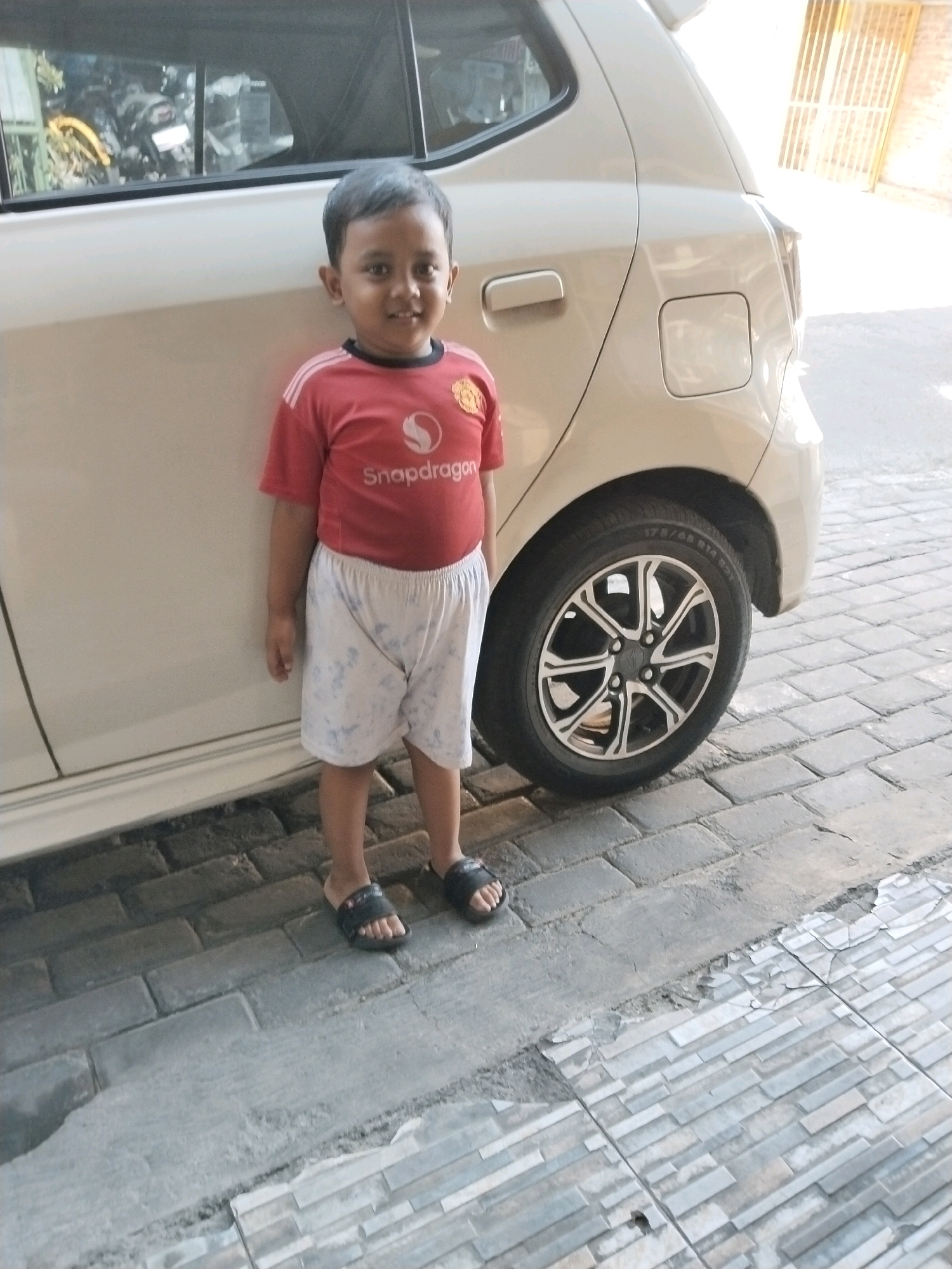 A young boy stands in front of a light-colored car on a paved outdoor surface. The boy, who appears to be of East Asian descent, is wearing a red t-shirt with a white logo and white shorts with a light blue pattern. He is also wearing black sandals. The car is a hatchback, and its rear wheel and fender are visible. The ground is paved with interlocking brick-like stones and some concrete sections. In the background, a glimpse of a building with a yellow gate and a brick wall suggests an urban or suburban environment. The lighting suggests it is daytime, with sunlight casting shadows. The image seems to have been taken in Pepelegi, Indonesia, an area characterized by its residential and commercial developments.