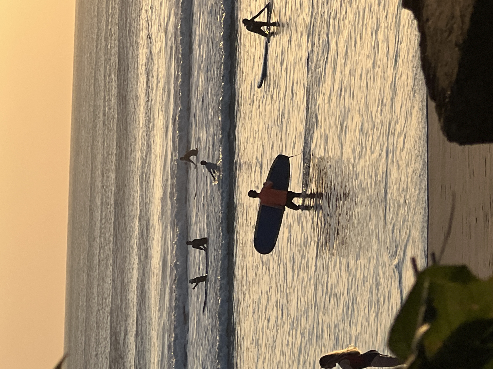This image depicts a group of surfers in the ocean at Kuta Beach, Indonesia, during sunset. The sky is a warm orange hue, casting a golden glow over the water. Several individuals are visible in the water, some on surfboards and others wading. In the foreground, a person is carrying a dark blue surfboard and walking into the shallow water. Further out, other surfers are positioned on their boards, with some appearing to be paddling or waiting for waves. The ocean is calm with gentle waves breaking near the shore. The beach in the lower portion of the image is sandy and appears to be empty except for some foliage in the far left foreground and a dark rock formation in the right foreground. The overall scene suggests a serene and active coastal environment during the evening.