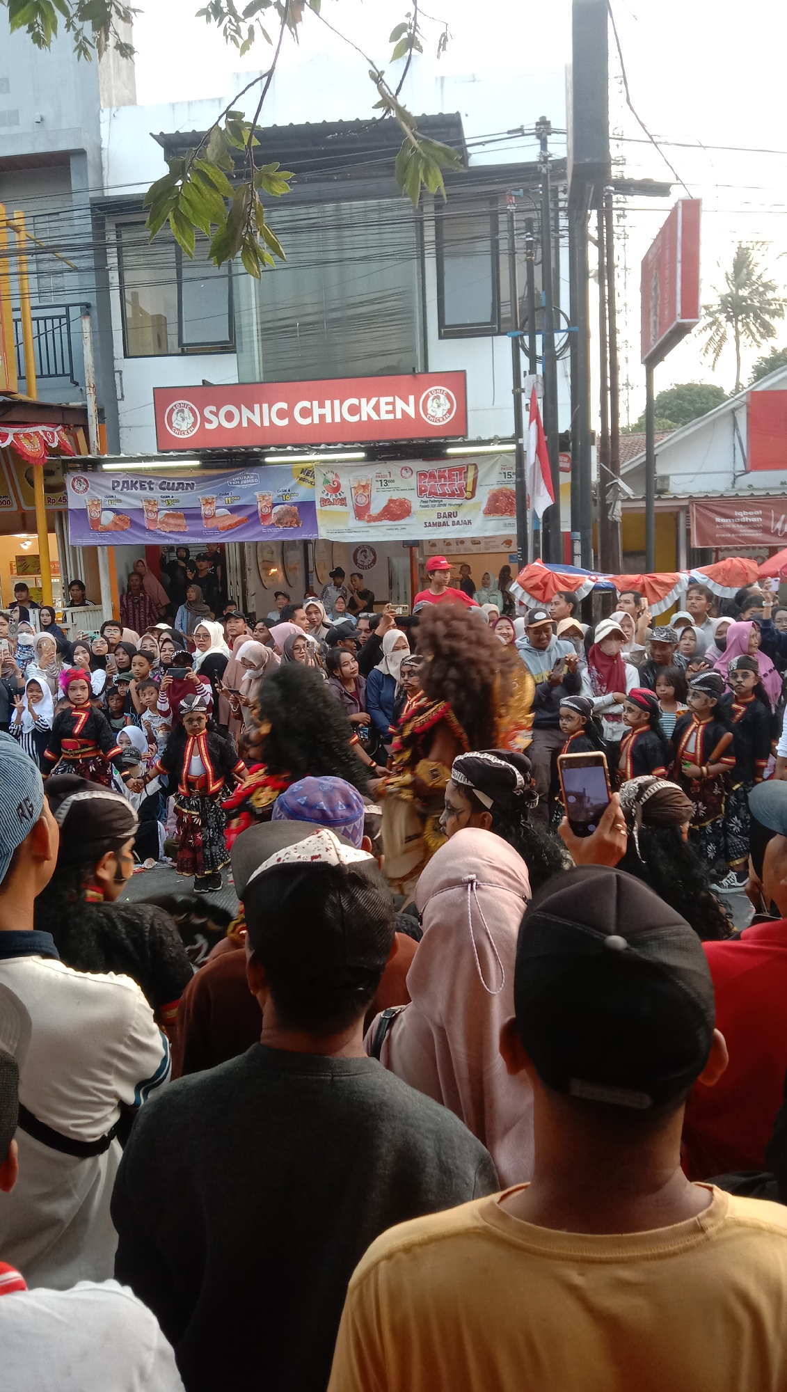 The image depicts a lively outdoor scene, possibly a street festival or performance, in Jatisawit, Indonesia. A crowd of people, including children and adults, are gathered to watch a performance. In the foreground, several individuals are observing the event, some are facing away from the camera. In the midground, performers in traditional attire, possibly part of a cultural dance or parade, are the central focus. One performer wears a distinctive, large, dark, bushy wig and elaborate costume, while others, including children, are dressed in matching outfits with black, red, and gold accents, some with face paint. Behind the performers, banners and signs indicate commercial establishments, including "SONIC CHICKEN" prominently displayed on a building, suggesting a commercial area. The environment appears to be an urban or semi-urban setting with buildings, power lines, and street decorations visible. The overall atmosphere suggests a community event with cultural elements.