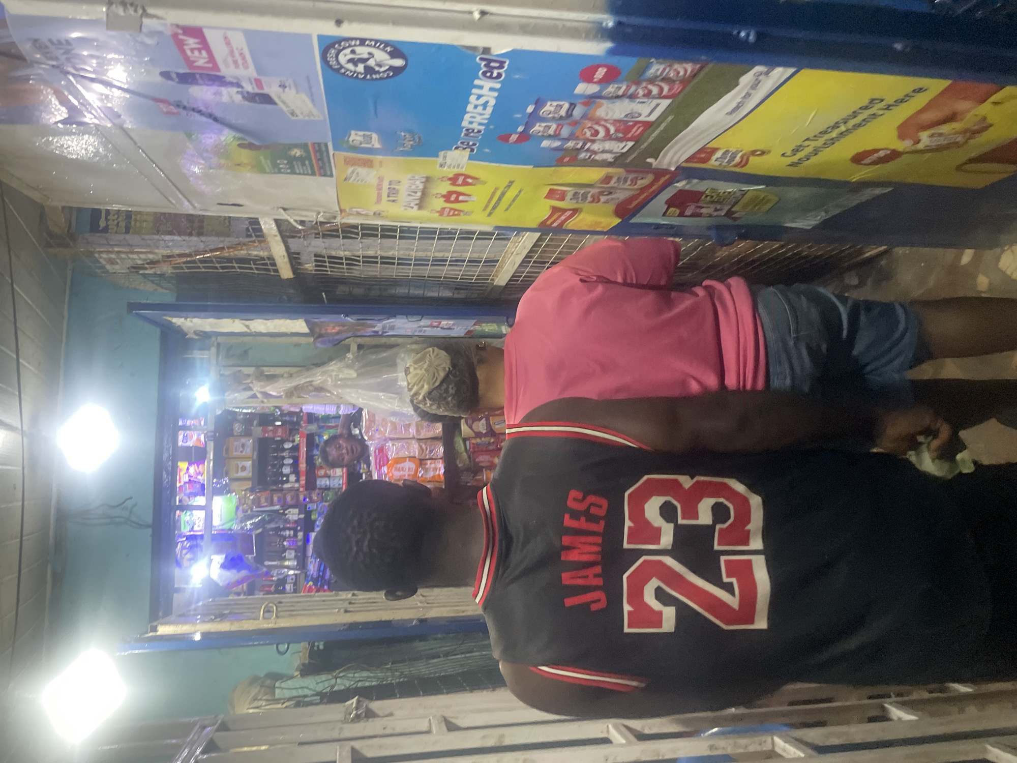 The image depicts an indoor scene, possibly a small shop or market stall, located in Du, Nigeria. Two individuals are visible from behind, facing away from the camera and towards a store interior. The person in the foreground is wearing a black sleeveless jersey with "JAMES" and the number "23" in red lettering. The second person, slightly behind and to the right, is wearing a pink t-shirt and denim shorts.
The store interior is filled with shelves stocked with various products, including bottled drinks and packaged goods. Lighting fixtures are visible on the ceiling, providing illumination. The exterior environment shows a narrow passageway, with metal grating and blue vertical posts on the left. Advertisements and posters are displayed on the wall to the right. The overall impression is of a bustling, indoor commercial space.