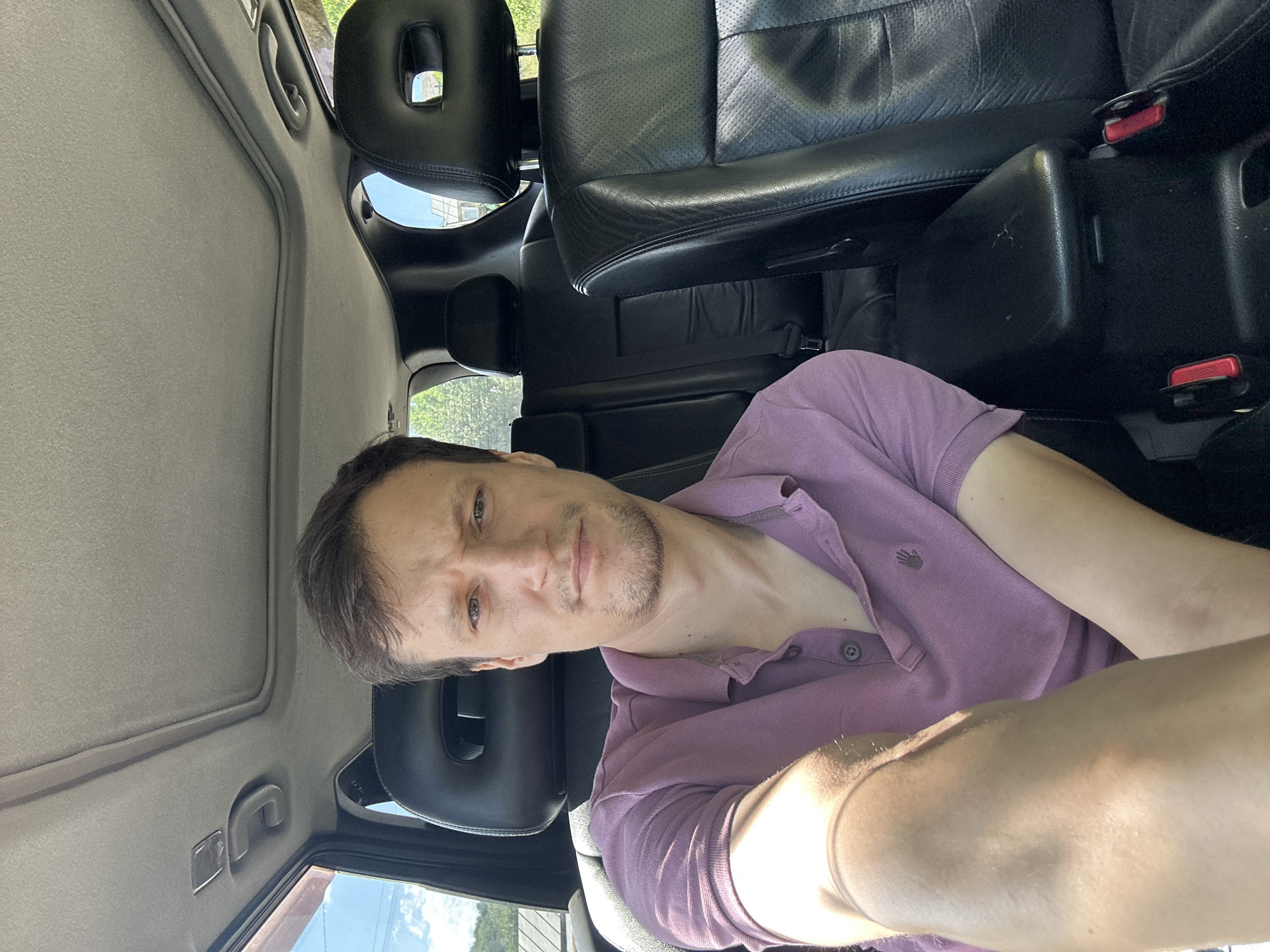 A man is taking a selfie inside a car, seated in the back. He is wearing a purple polo shirt and has short brown hair and stubble. The car's interior is visible, with black leather seats, headrests with cutouts, and a sunroof. The background shows a slightly blurred outdoor scene with greenery and buildings. The image appears to be taken during the day, with sunlight filtering into the car. The man is looking directly at the camera with a neutral expression.