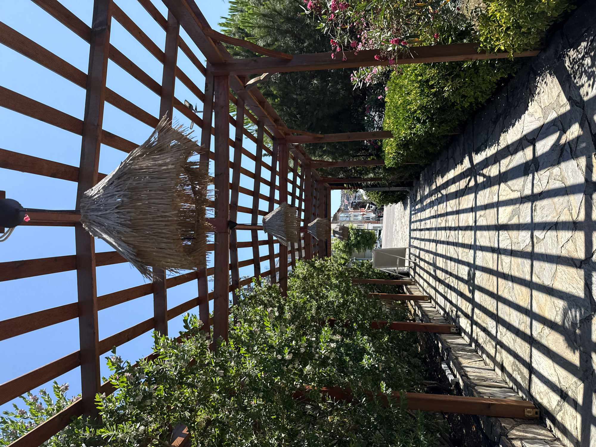 The image shows a pathway under a wooden pergola in Şile, Turkey. The pergola has a slatted roof, and straw lamps hang from it. Lush green trees and flowering bushes line the pathway. The ground is paved with irregular stones, and the sunlight casts a pattern of shadows from the pergola onto the ground. In the distance, a street with buildings is visible. The sky is clear and blue. There are no people or animals in the image.