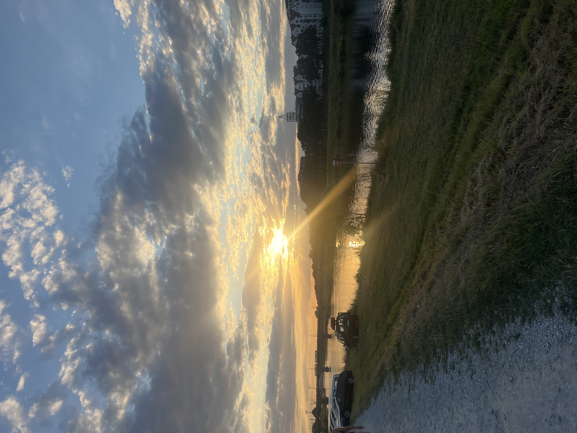 The image depicts a sunset over the Oder River in Wrocław, Poland, likely viewed from the embankment on the west bank. The sun, a bright orb, is partially obscured by dramatic clouds, casting a warm, golden light across the sky and reflecting on the water's surface. In the foreground, a gravel path runs along the left side of the image, with a grassy embankment sloping down to the river. Several parked cars are visible on the grass near the path, including a dark SUV with a roof rack. Across the river, the cityscape of Wrocław is silhouetted against the colorful sky. Buildings with white facades and red roofs line the riverbank, with a prominent tower-like structure visible in the distance. A bridge spans the river further in the background, with what appears to be a train or tram crossing it. The overall scene conveys a peaceful and picturesque evening ambiance.