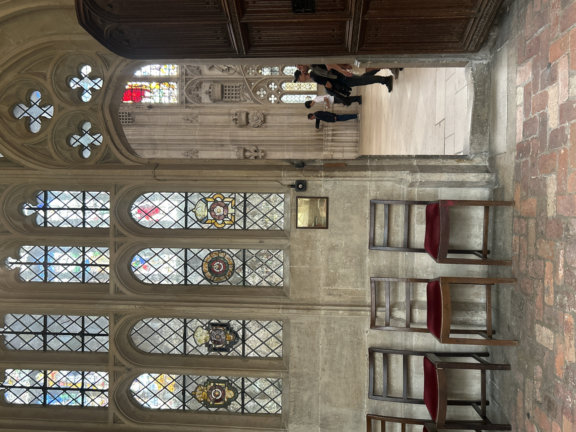 The image captures an interior view of a grand building, likely a cathedral or historic college hall, in Cambridge, United Kingdom. The scene is characterized by Gothic architecture, prominently featuring large stained-glass windows with intricate diamond-patterned leadwork and colorful depictions. The walls are constructed from large, rough-hewn stone blocks, contributing to the ancient ambiance.
In the foreground, to the left, a row of four dark wooden chairs with red velvet seats are lined up against the stone wall. These chairs appear to be simple in design, likely for visitors or congregants.
To the right, a large, ornate wooden door stands ajar, revealing a glimpse into an adjacent hallway or chamber. Beyond this doorway, several individuals are visible. A man in a dark jacket and light pants is pointing towards something in the distance. Two other men are standing near the entrance, looking towards the interior. One is wearing a black cap and a dark t-shirt, holding a jacket over his arm, while the other is wearing a dark t-shirt and lighter colored pants.
The floor in the foreground is paved with red brick in a herringbone pattern, transitioning to lighter stone tiles further into the building. The overall lighting suggests an overcast day or an interior space with limited natural light. The architecture, particularly the stained glass and stonework, strongly indicates a location within Cambridge, possibly within one of its famous colleges or religious institutions.