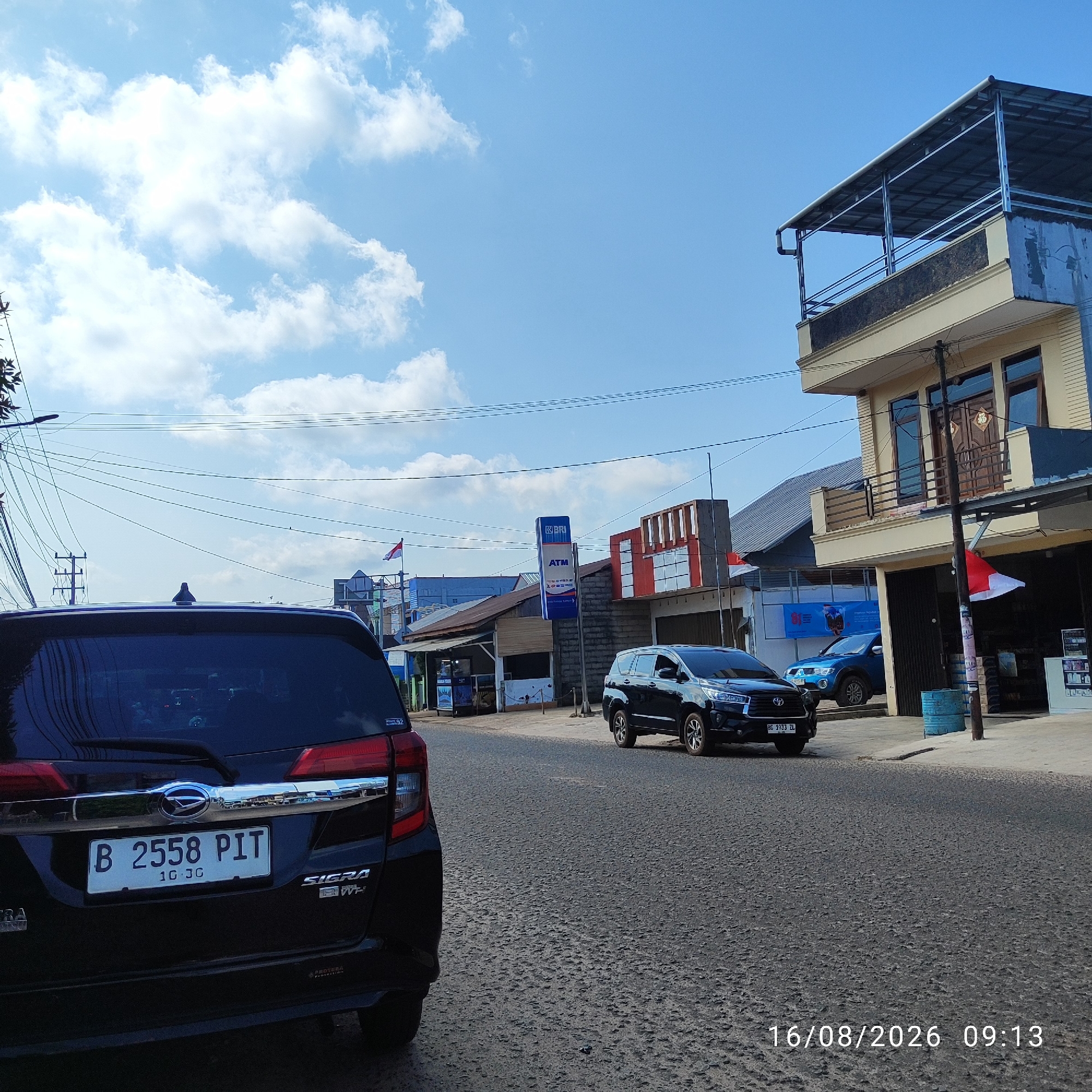 The image depicts a street scene in Pembaharuan, Indonesia, on August 16, 2026, at 09:13. The sky is blue with scattered white clouds. In the foreground, the rear of a black car, identified by its license plate "B 2558 PIT", is visible. The car is parked on a paved road. To the right of the black car, a dark-colored SUV is also present on the road. Further down the street, on the right side, is a multi-story building with a second floor featuring a balcony. Red and white flags, likely the Indonesian flag, are displayed on the building and nearby structures. Several other buildings line the street, some appearing to be shops or small businesses, with awnings and storefronts. An ATM sign for "BRI" is visible on one of the buildings. Overhead, a complex network of electrical wires crisscrosses the scene. The environment suggests an urban or semi-urban setting with modest commercial establishments. No people are clearly visible in the image.