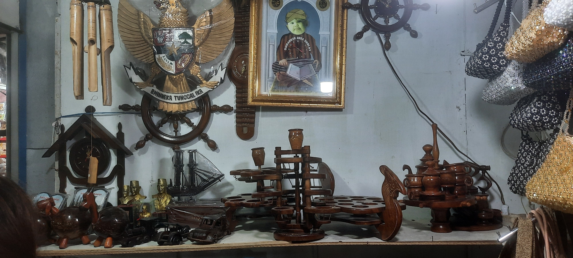 The image depicts a cluttered stall or shop interior, likely in Blitar, Indonesia, showcasing a variety of handcrafted items. Dominating the left side of the frame is a large wooden Garuda symbol, the national emblem of Indonesia, with the motto "BHINNEKA TUNGGAL IKA" visible beneath it. To the right of the Garuda is a framed portrait of a child wearing traditional attire and holding a book. Various wooden decorative items are displayed, including a ship model, several small carved animals, and what appear to be ornate wooden racks holding small containers, possibly for tea or spices. Several beaded bags, in black, white, and gold, hang from a rail on the right side of the image, suggesting they are also for sale. A wooden clock is mounted on the wall above the Garuda. The lighting suggests it is daytime, with the stall appearing to be indoors, possibly an open-air market stall or a shop. No people are clearly visible in the scene, though a sliver of someone's dark hair is present at the bottom left corner. The overall impression is of a vibrant, artisanal marketplace.