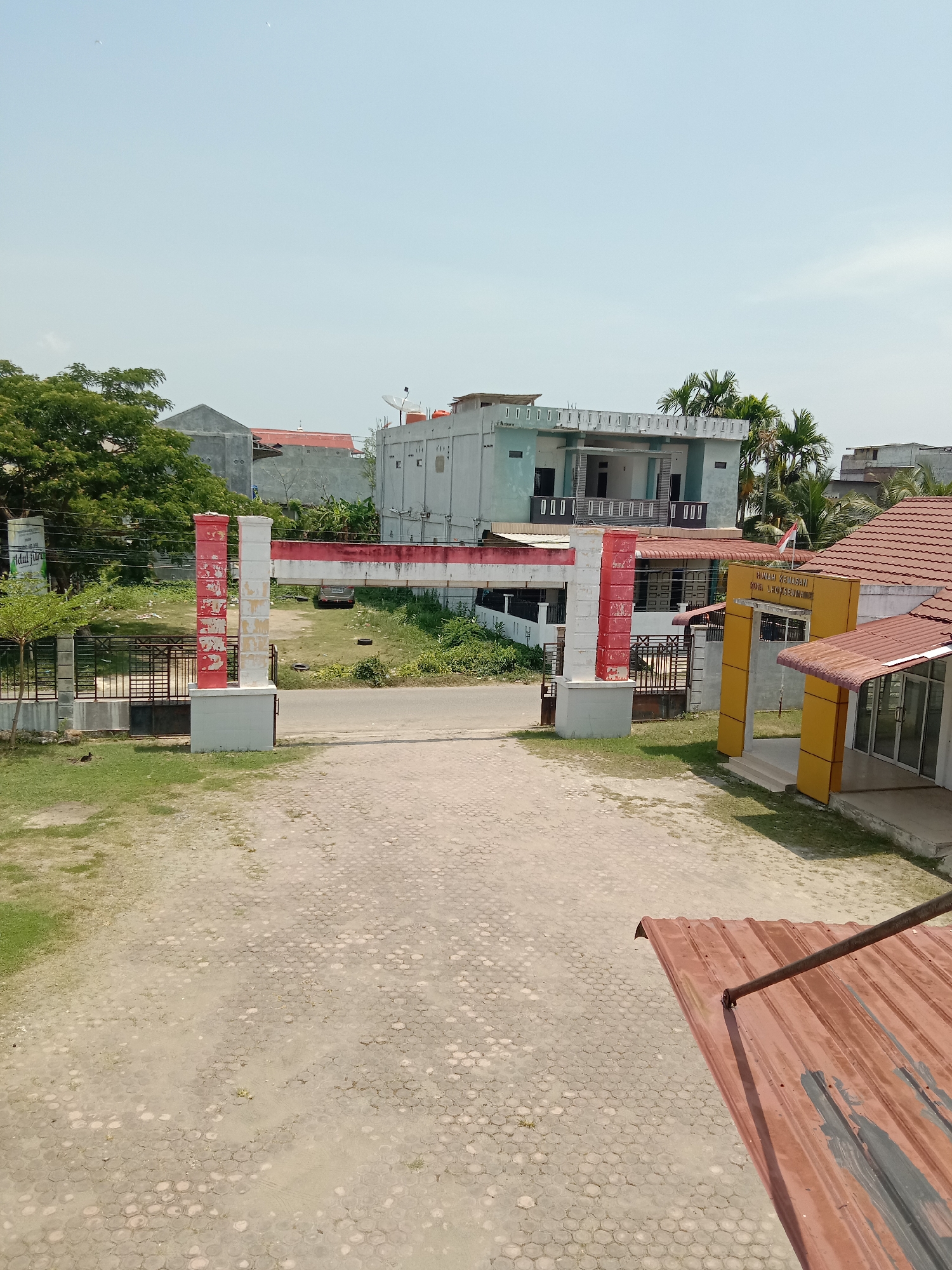 The image presents an outdoor scene under a bright, clear blue sky, indicating daytime conditions. The perspective is slightly elevated, looking down into an entrance area in Lhokseumawe, Indonesia.
In the foreground, a section of a reddish-brown corrugated metal roof is visible in the bottom right corner. The ground consists of patterned concrete pavers, mostly dirt-colored, with scattered patches of green grass.
A prominent red and white gate structure, echoing the colors of the Indonesian flag, marks the entry point. This gate spans a paved road leading into the complex. To the left of the gate, a black metal fence borders a grassy area with mature green trees. To the right, a yellow-painted building entrance with a brown-tiled roof is visible. The text "RUMAH KEMASAN KOTA LHOKSEUMAWE" is clearly displayed on the archway of this yellow building, and an Indonesian flag flies from a pole nearby.
Beyond the gate, a light blue two-story building with a flat roof and balcony railings stands on the left, with other residential-style buildings visible further back. On the right, more of the complex's buildings with red-tiled roofs are seen, alongside lush green palm trees. A partially obscured white sign with "Abdul Jabar" or similar text is faintly visible to the left of the road, before the main gate. The scene depicts a functional, likely public or institutional facility entrance in a semi-urban setting.