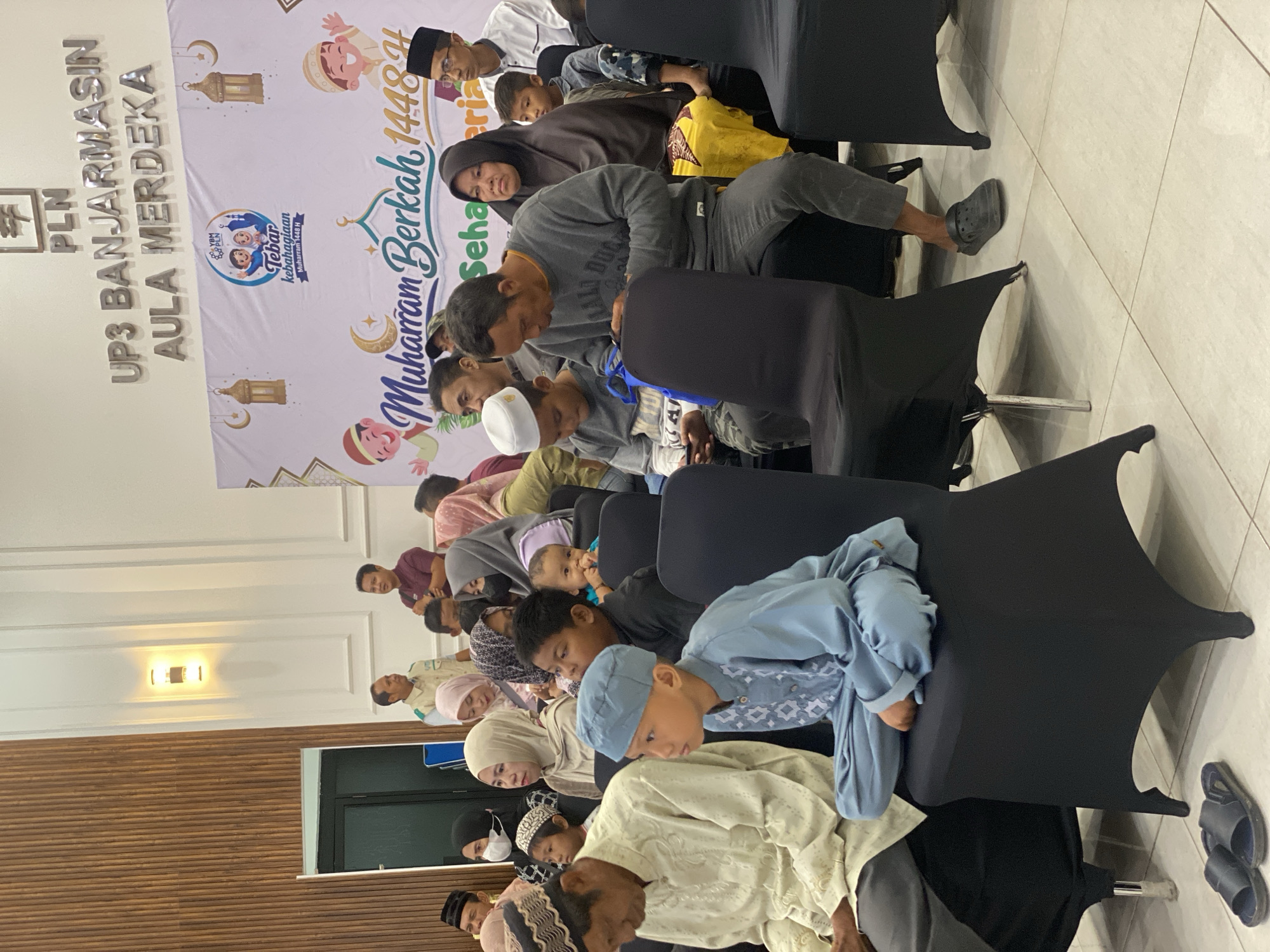The image depicts an indoor gathering at the PLN UP3 Banjarmasin Aula Merdeka in Banjarmasin, Indonesia, likely for a Muharram 1448 H celebration. A diverse group of people, including men, women, and children, are seated in black-covered chairs facing forward, suggesting they are attending an event or presentation. The backdrop features a banner with text related to the celebration and decorative elements like lanterns and crescent moons, indicative of an Islamic festive occasion. The lighting appears to be artificial and ambient. Several individuals are dressed in traditional Islamic attire, such as peci hats and headscarves, adding to the cultural context of the event. The overall atmosphere seems to be one of communal gathering and shared celebration.