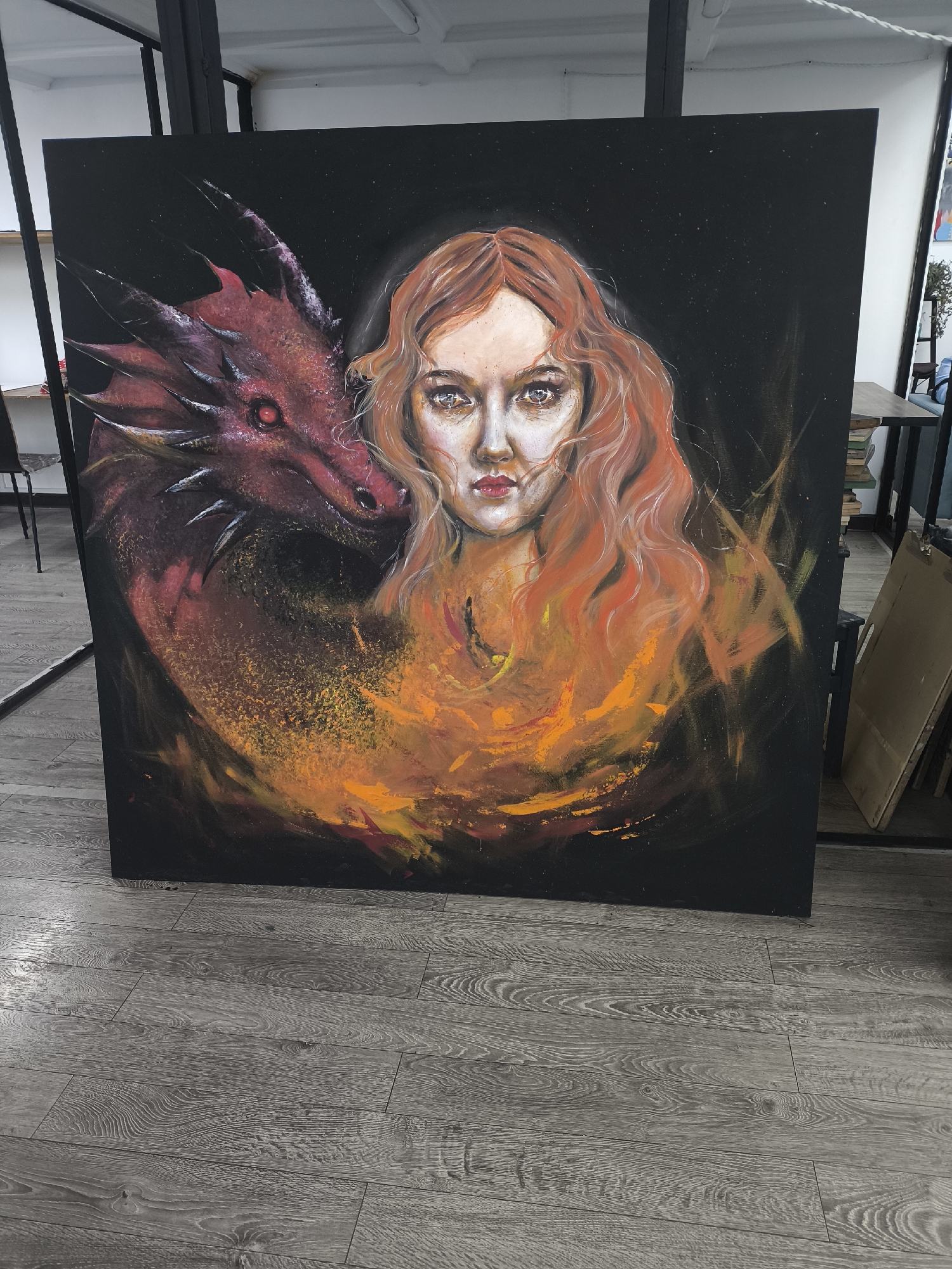 The image shows a large painting leaning against a wall with gray wooden flooring. The painting features a woman with auburn hair and a red dragon. The background of the painting is black, and orange and yellow hues surround the woman and dragon, suggesting fire. The painting is placed in what appears to be an art studio or workshop, based on the scaffolding in the background and the presence of other canvases or materials. The exact location cannot be determined, and the information about Moscow, Russia is not discernible from the image.