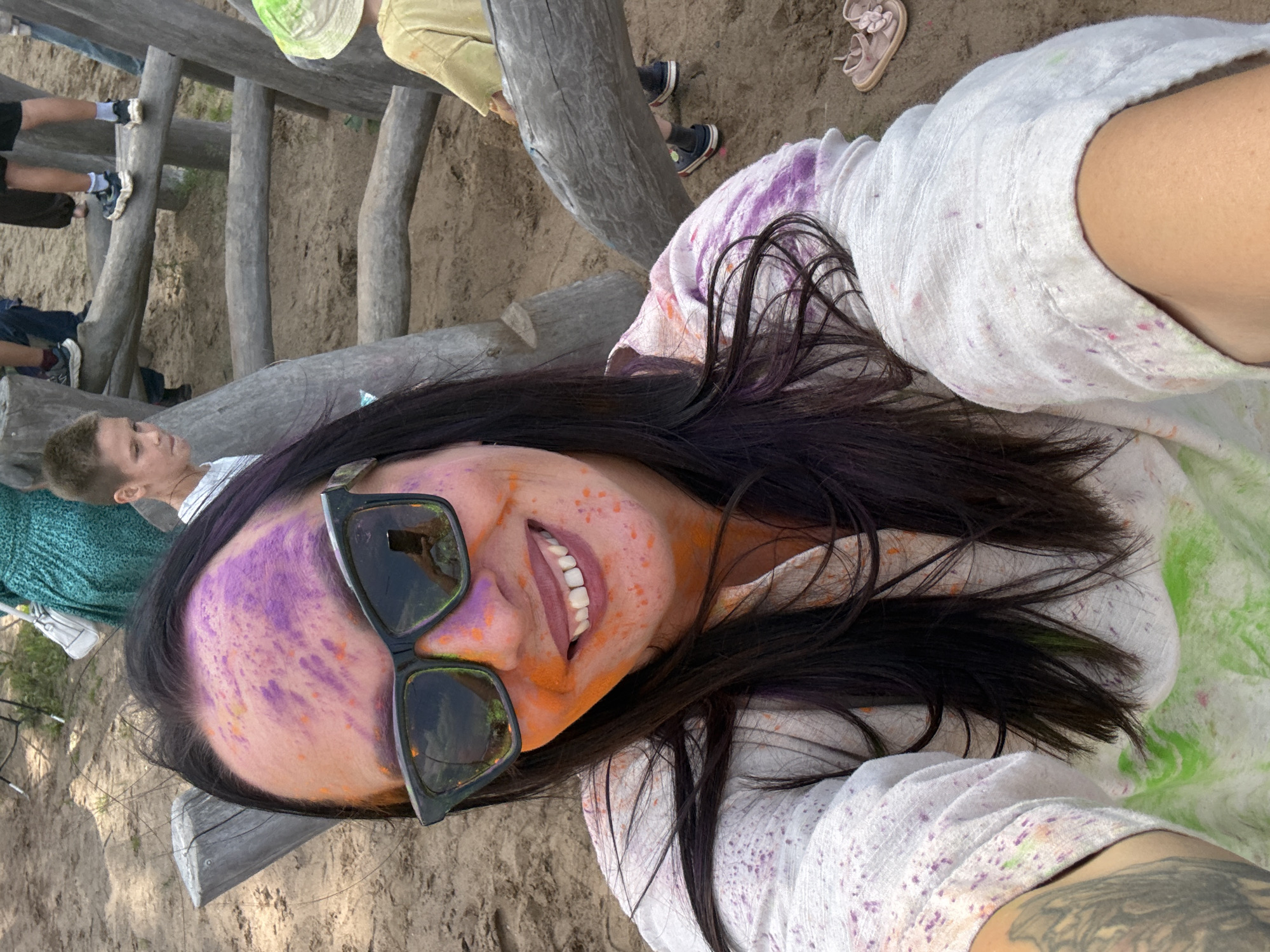 An outdoor, close-up selfie shows a woman with purple and orange powder pigment on her face and white clothing, smiling with her mouth open. She is wearing dark sunglasses. In the background, there is a wooden structure made of logs, sand, and several other people, including two children. The woman's arm on the left side of the image is visible, showing a tattoo. The setting appears to be a park or recreational area. It is not possible to determine the specific location in the city of Chaikovsky, Russia from the provided image.