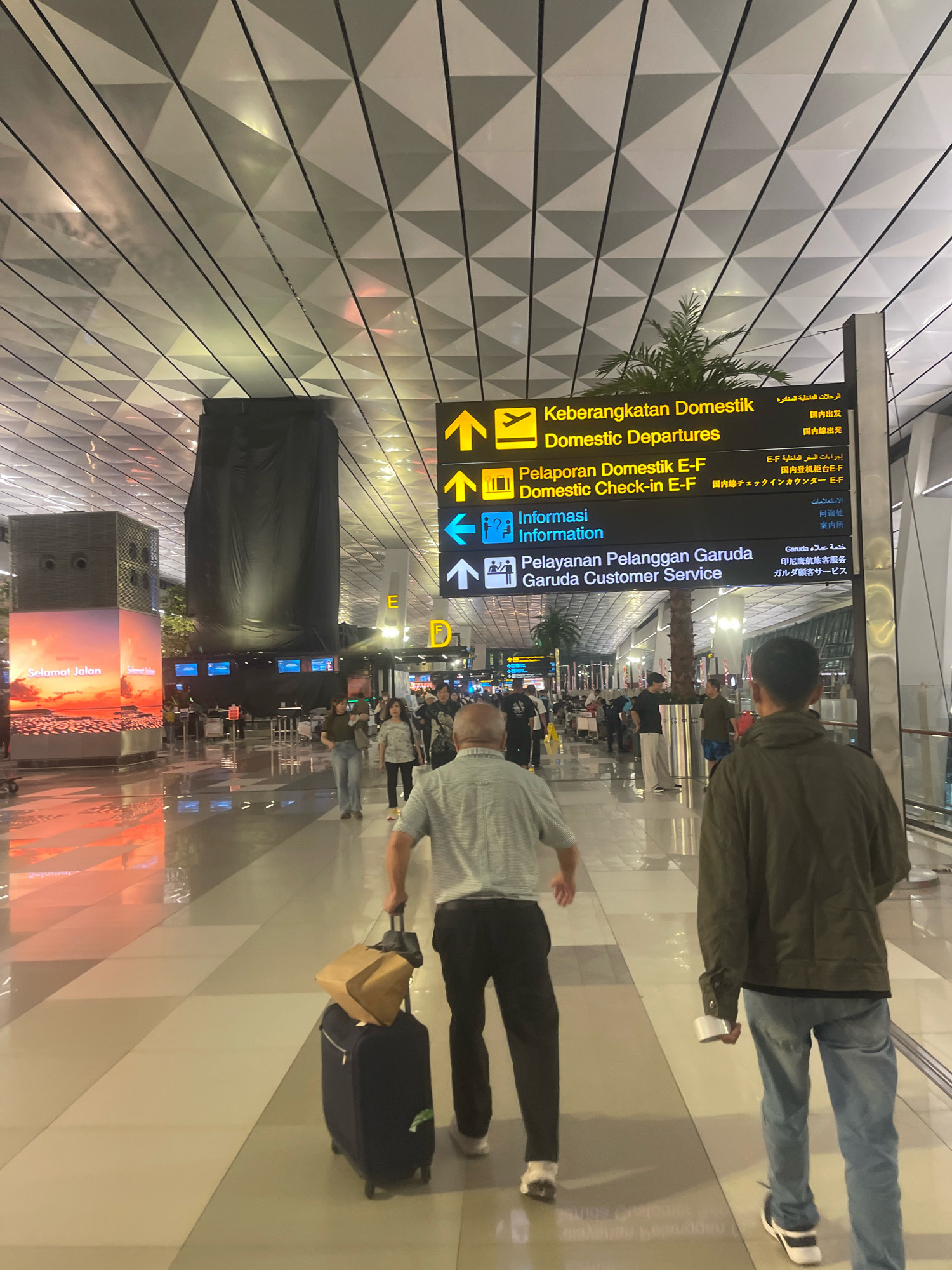 The image depicts an interior view of an airport terminal. In the foreground, two men are walking away from the camera. The man on the left is wearing a light blue and white striped shirt and dark pants, and is pulling a dark blue suitcase with a brown bag on top. The man on the right is wearing a green jacket and blue jeans.
In the midground, several other people are walking in the same direction as the foreground subjects. There are also signs with directional arrows and text in multiple languages, indicating airport services such as "Domestic Departures," "Domestic Check-in," and "Garuda Customer Service." A large black tarp covers a cylindrical structure. To the left, a digital billboard displays the text "Selamat Jalan."
The environment is a modern airport terminal with a tiled floor and a geometric ceiling. The lighting is artificial and reflects off the polished surfaces. The location is specified as Pajang, Indonesia.