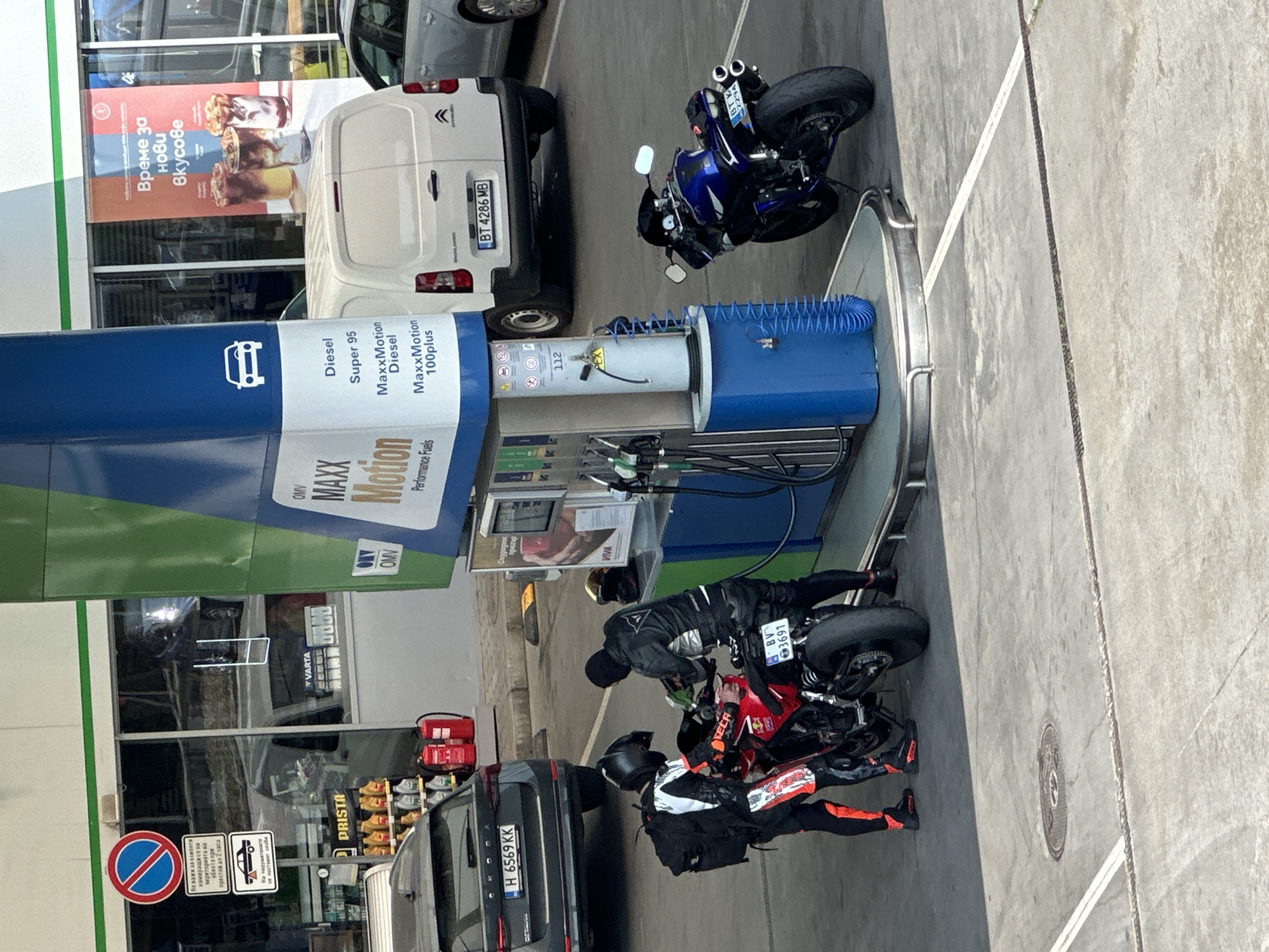 In Veliko Tarnovo, Bulgaria, two individuals in motorcycle gear are at a gas station. One person is seated on a red motorcycle, wearing black and orange riding suits, while the other stands next to them, leaning towards the fuel pump. A blue and white fuel dispenser with "OMV MAXX Motion" and "Performance Fuels" visible on its side is the central object. Next to it, a blue hose is coiled. A dark grey car with license plate "H 6569 KK" is parked nearby. Another motorcycle, blue and black, is parked to the right of the fuel dispenser. In the background, a white van with license plate "BT 4286 MB" is visible, along with advertisements for "VIVA" and coffee. Signage in Bulgarian indicates parking regulations and a "DrISta" brand. The scene appears to be during daylight, with no specific weather indicators. The overall impression is a common stop at a gas station for motorcyclists.