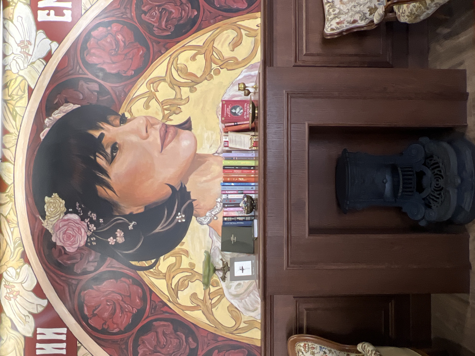 This image captures a richly decorated interior, likely a library, study, or a room with a strong artistic theme, consistent with the elegance of Saint Petersburg, Russia. The primary focus is a large, ornate wall painting on the left, depicting the head and shoulders of a young woman in profile, looking upwards and to the right. She has dark hair styled with delicate white and pink roses and tiny white blossoms. Her skin is fair, and her expression is serene or contemplative.
The woman's portrait is set within a circular or arched frame, edged in gold, which features elaborate golden scrollwork against a lighter background. Surrounding this frame are smaller, dark circular insets containing red roses, and larger white lily-like flowers are visible in the background, particularly on the far left. The painting style is realistic for the woman, combined with decorative Art Nouveau-like motifs for the surrounding elements.
To the right of the painting is a dark brown wooden structure, possibly a built-in cabinet or shelving unit, with clearly defined panels and a sculpted ledge. Within a niche or on shelves, a collection of books is neatly stacked, both vertically and horizontally. Many of the books have colorful spines. Several decorative items are interspersed among the books, including a small, gold-toned chalice-like object, a green glass dish or ashtray on a stand, and a small framed picture displaying a simple sketch or print, possibly of an airplane.
In a lower, larger recess within the wooden structure, a distinctive, dark cast-iron object rests. It appears to be an antique decorative piece, possibly a part of an old stove, fireplace insert, or an elaborate stand, characterized by intricate, industrial-looking details and a round, coiled motif. Partially visible upholstered furniture, likely a chair or armchair with a patterned fabric, can be seen at the top and bottom edges of the image, suggesting comfortable seating within the room.
Visible text on the painting's left edge includes partial white Cyrillic letters, possibly "ЕН" or "КИ". On one of the red books, Cyrillic text can be deciphered as "Лев Толстой" (Lev Tolstoy), suggesting classic Russian literature. The scene is static, devoid of human activity, and illuminated by what appears to be ambient indoor lighting, enhancing the warm and inviting tones of the decor.
