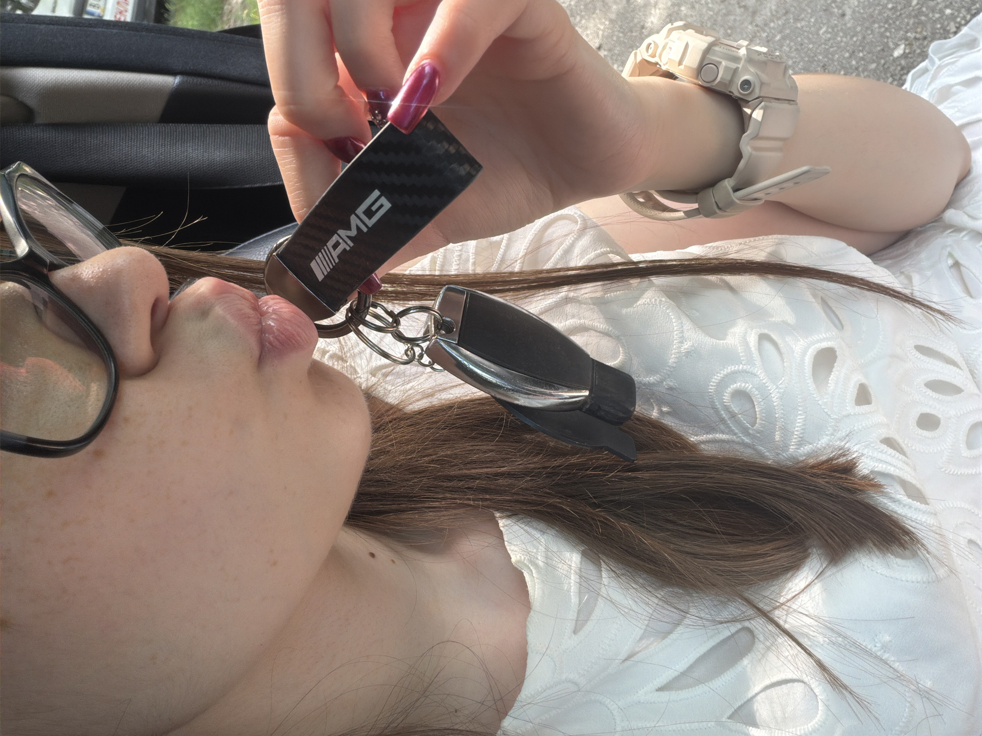 A close-up outdoor portrait features a female individual with long brown hair, fair skin, and visible freckles on her face and neck. She wears dark-rimmed glasses and a white top with an intricate cut-out or embroidered pattern. Her lips are puckered in a kissing gesture, directed towards a black rectangular keychain with a carbon fiber texture and the white "AMG" logo. This keychain is attached to a keyring, alongside a modern black automotive key fob. Her right hand, with glossy dark red/maroon manicured nails, holds the keys. On her right wrist, she sports a light beige/tan digital wristwatch, resembling a G-Shock style. The background is blurred, showing green foliage, a dark seatbelt, and other elements suggestive of a car interior. A partially visible license plate in the upper right background displays the regional code "BA," confirming the location as Varna, Bulgaria.