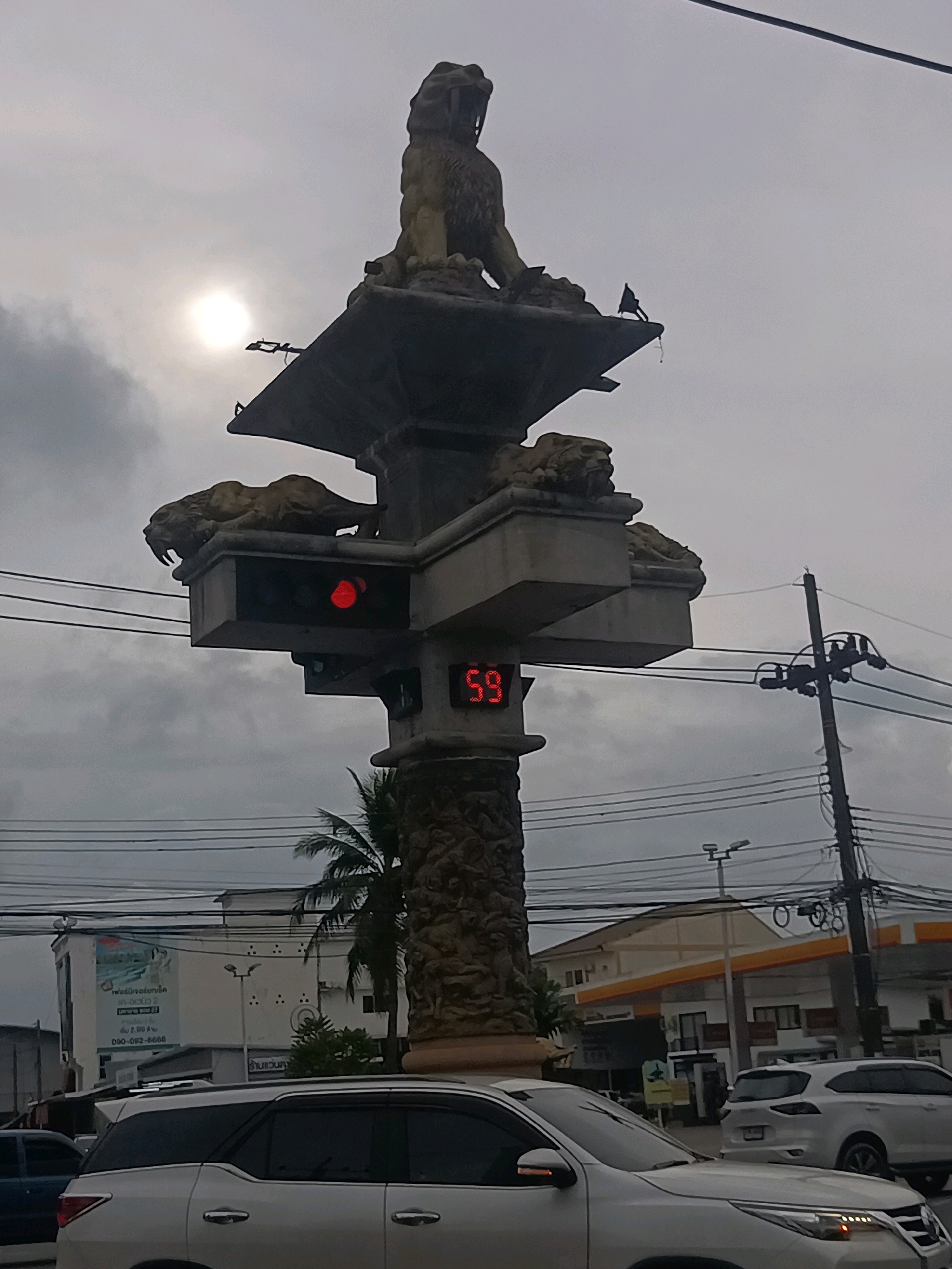 The image captures a traffic intersection in Tambon Pak Nam, Thailand, under an overcast sky. Dominating the center is a traffic signal mounted on an ornate pillar decorated with bas-relief carvings and topped with sculptures of lions. A red traffic light is illuminated, and a digital display shows "59," likely indicating seconds remaining. A white SUV is prominently featured in the foreground, partially obscuring the pillar and other vehicles behind it. In the background, a building with a prominent sign, possibly a business or residential complex, and a gas station with a yellow and orange canopy are visible. Electrical power lines crisscross the scene, supported by wooden poles. The overall impression is of a typical urban street scene in Thailand.