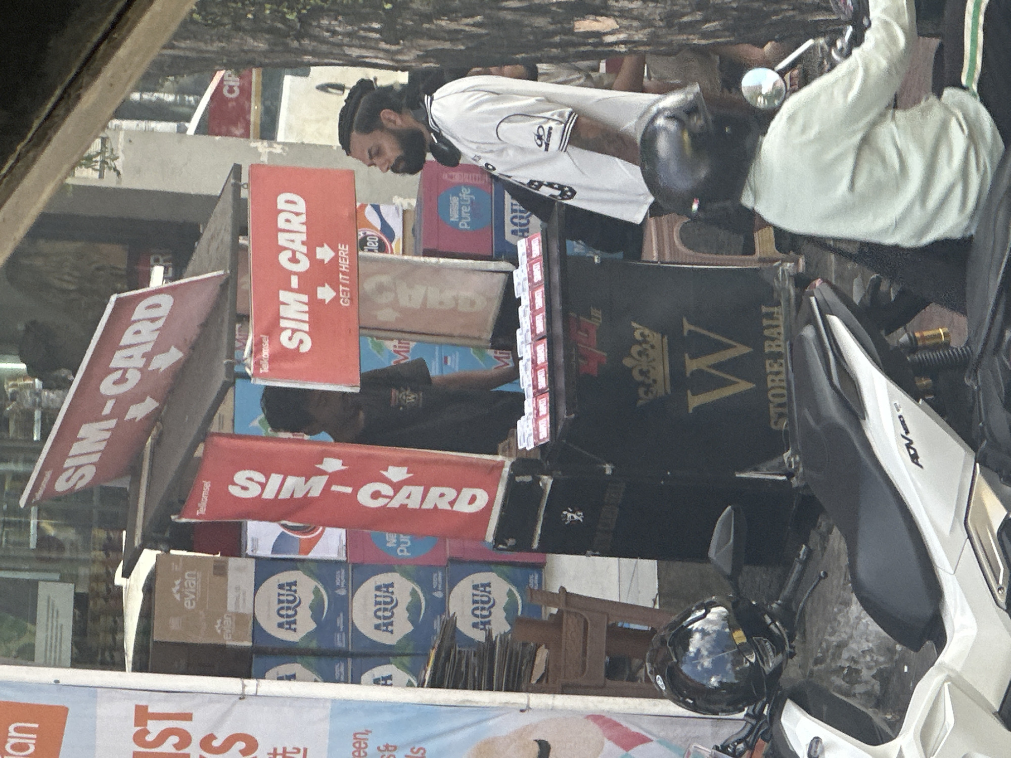 A man with dark, braided hair and a beard stands at a street-side stall in Ubud District, Indonesia, selling SIM cards. He wears a white t-shirt with black trim and a black backpack. The stall is cluttered with stacks of bottled water, notably "AQUA" and "Nestle Pure Life," and features red banners advertising "SIM-CARD" with arrows pointing downwards. Another man, partially visible in the background, wears a black t-shirt. The scene is framed by parked motorcycles, one of which has a black helmet resting on its handlebars, reflecting the sky. The stall is situated outdoors, suggesting a busy street environment.