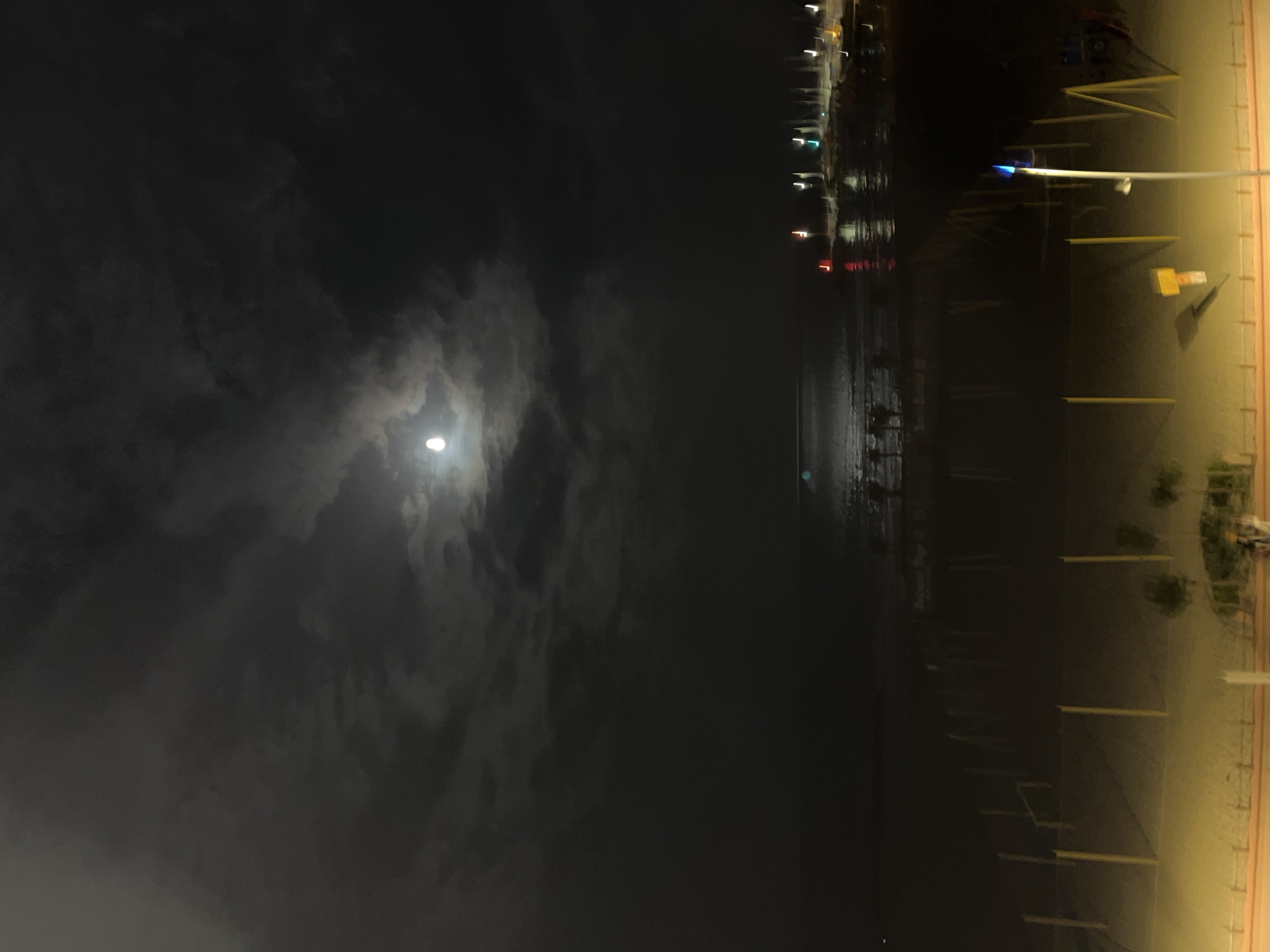 The image portrays a tranquil nighttime scene, likely at a coastal location such as Palavas-les-Flots, France. The dark sky dominates the upper left, where a luminous full moon shines through a patchy covering of soft, grey clouds, casting a diffused glow. The clouds range from thin veils to denser formations, partially obscuring the moon's bright disk.
Stretching across the middle of the frame is a calm body of water, possibly the Mediterranean Sea or a large lagoon. Its surface reflects a distant scattering of blurry, colorful artificial lights—including reds, greens, and whites—suggesting structures like a distant town, a pier, or a promenade along the opposite shore. These reflections create shimmering trails on the water.
To the right, a sandy beach or ground area is visible, illuminated by a warm, yellow-orange light, likely from unseen artificial sources. Several thin, dark poles are regularly spaced vertically within this sandy expanse, possibly marking a boundary or part of a boardwalk structure. A prominent light pole, topped with a distinctive blue light, stands out in the mid-right portion of this illuminated area. Faint outlines of vegetation are also discernible near the beach.
The overall atmosphere is one of serene solitude, with no people or specific activities taking place. The time of day is clearly night, characterized by the dark sky, moon, and artificial lights. The presence of water, beach, and reflections strongly indicates a waterfront setting. No text is visible in the image.