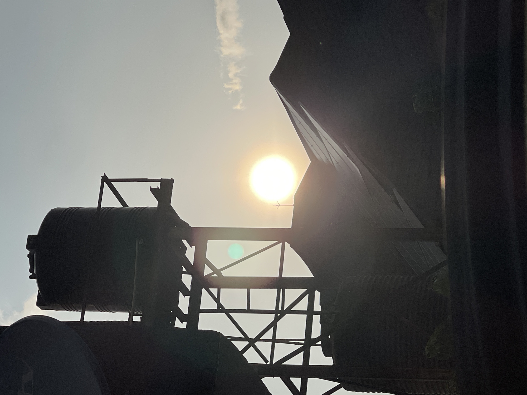 This is a silhouetted, vertical, outdoor, medium shot of a water tower and the roof of a building against a bright, hazy sky with the sun partially visible. The scene is likely in Abuja, Nigeria, given the style of the water tank and the construction of the roof. The water tank is tall and cylindrical with ribbed sides, supported by a dark, metal frame structure. Below it, another, wider tank is visible, also in silhouette. The roof of the building is dark and angular, with a sharp peak. The sun is a bright, white circle, its light creating a lens flare and casting a green circular reflection on the metal structure. A thin cloud formation is visible in the upper right portion of the sky. The foreground is dominated by a dark, horizontal surface. No people are visible in the frame.
