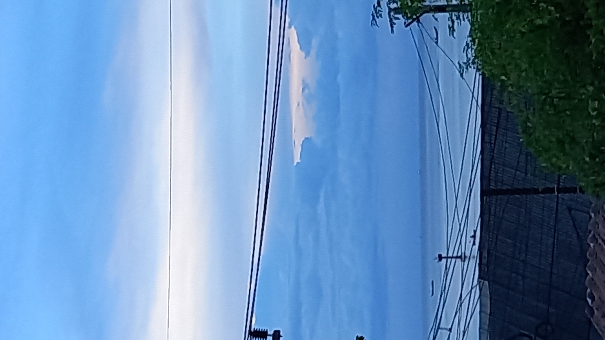 The image depicts a partially rotated view of a sky with clouds, power lines, and some foliage on the right side. The sky is predominantly blue with patches of white clouds, suggesting it might be daytime. The presence of power lines indicates an urban or semi-urban setting. On the far right, there is lush green foliage and a dark structure that appears to be a fence or a wall, partially covered by a roof-like element at the bottom. There are no people visible in the frame. The overall scene is tranquil, with the focus on the interplay of light and shadow in the sky and against the infrastructure. The location context provided is Koh Phangan, Thailand.
