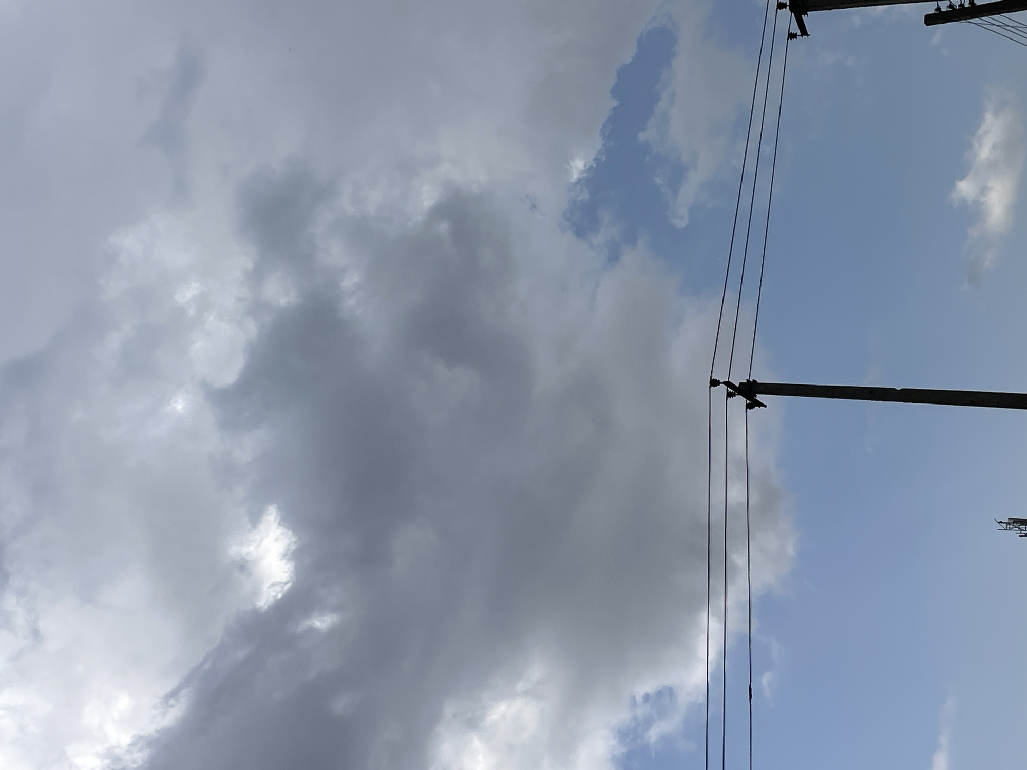 The image shows a sky with a mix of grey and white clouds on the left side and a clear blue sky with scattered white clouds on the right. A utility pole with wires runs vertically through the right half of the image, with two horizontal poles visible at the top and bottom of this section. The wires are dark and run parallel to each other. The scene appears to be outdoors, with the sky suggesting daytime. The clouds indicate potentially changeable weather, perhaps a transition from overcast to clearer conditions. No people are visible, and the focus is on the sky and the utility infrastructure. The overall impression is that of an ordinary sky observation, possibly in a rural or semi-urban setting given the presence of overhead wires. There is no visible text in the image.