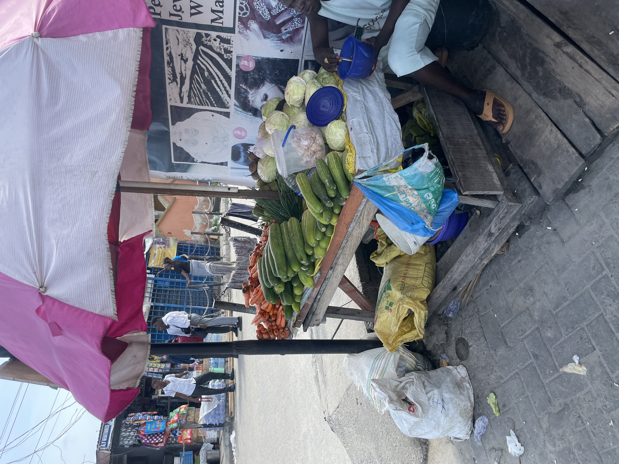 **Location:** Ado, Nigeria. Likely an outdoor market or street-side stall.
**People:** One person is visible. The individual is seated, wearing a light-colored top and shorts, and sandals. They are holding a blue container and appear to be interacting with vegetables on display. Other people walk in the background.
**Subjects:** A vendor or seller, vegetables for sale.
**Objects:**
* A vegetable stall constructed of wood.
* Pink and white striped umbrella providing shade for the stall.
* Various vegetables: carrots, cucumbers, cabbages. Some are stored in plastic containers or bags.
* Blue container with a lid.
* Several sacks on the ground, some yellow.
* Pictures, or posters on the stall wall.
* Various items for sale at a shop in the background.
**Actions:** The seated person is sorting, measuring, or handling vegetables.
**Interactions:** Interaction between the person and the vegetables is the only visible interaction.
**Environment:** Paved street. Shops and other street vendors visible in the background, suggesting a busy commercial area. The weather looks sunny and clear.