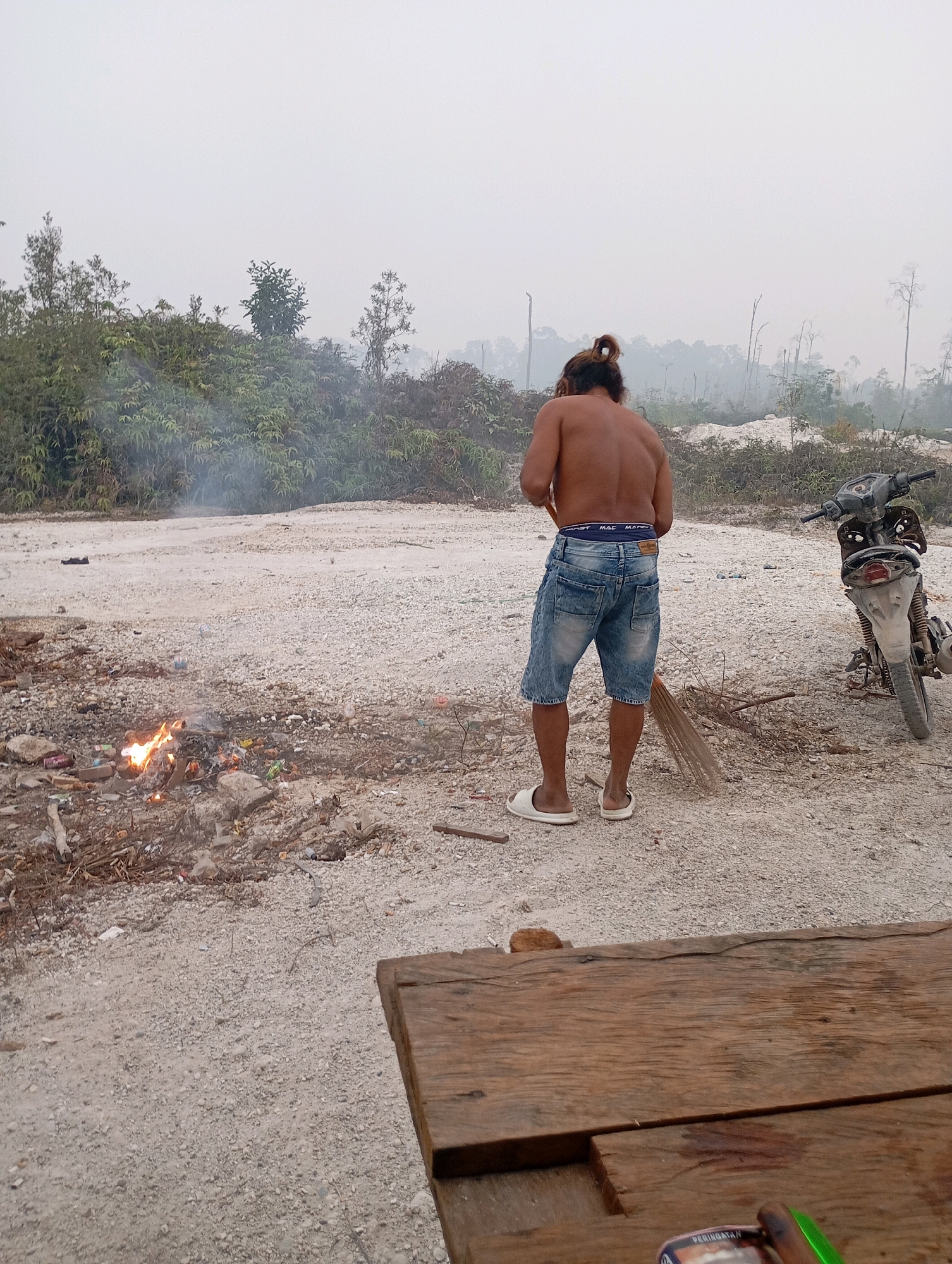 A shirtless man with his hair tied back in a bun is sweeping a dirt and gravel area with a broom. He is wearing denim shorts and white flip-flops. To his right, a dirt bike is parked. In the foreground, a small fire is burning amongst debris that includes cans and plastic items. The background shows dense vegetation and some scattered trees, with a hazy sky. The scene is located in Tumbang Tariak, Indonesia.