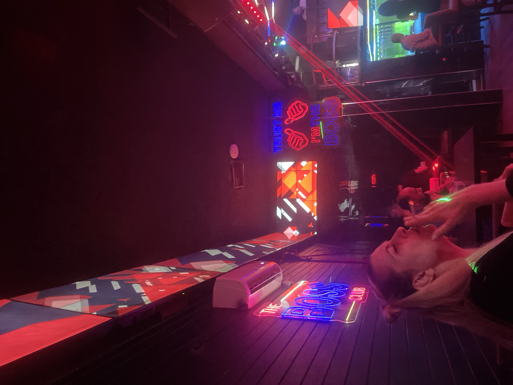 The image depicts the interior of a dimly lit establishment, possibly a nightclub or lounge, bathed in vibrant neon and red lighting. In the foreground, a woman with blonde hair tied back in a ponytail is seen in profile, exhaling smoke, suggesting she is smoking or vaping. She is wearing a black top. Behind her, the wall is adorned with a neon sign that reads "THE BOSS CLUB" in blue and pink lettering. Above the sign, an air conditioning unit is visible.
Further into the establishment, neon signs create a dynamic visual display. One sign reads "TRUST ME" in purple, with two neon thumbs-up icons above it. Below this, another neon sign says "I'M THE BOSS" in green and blue. The ceiling features a geometric pattern of red, blue, and white shapes.
In the background, several other patrons are present. A man in a light-colored striped shirt is seated at a bar or counter, facing away from the camera. Another individual, a woman with dark hair, is also seated. Tables and chairs are scattered throughout the space, some with red laser light beams crossing them. The overall atmosphere is characteristic of a nightlife venue. The specific location is identified as Amman, Jordan.