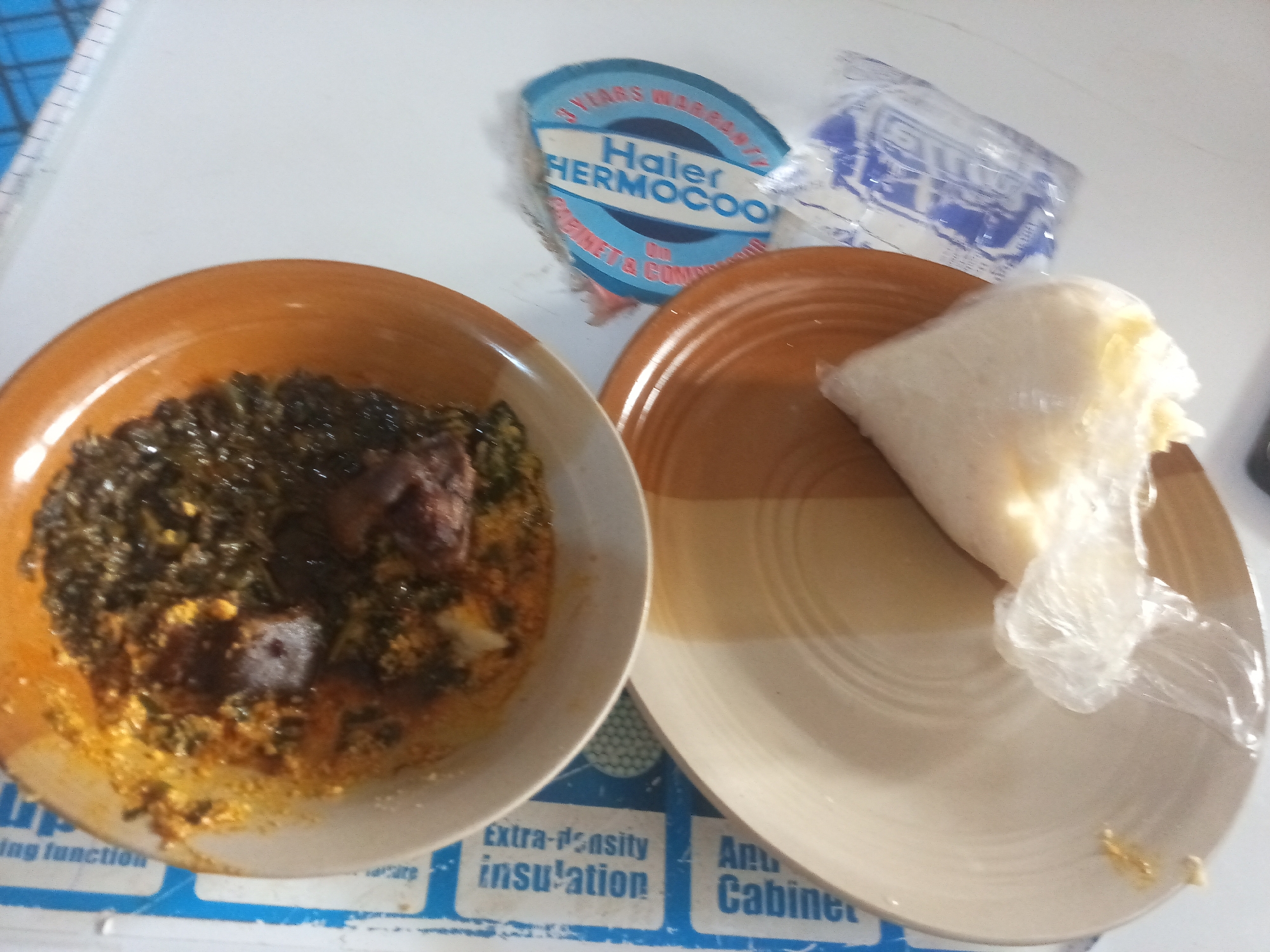 A low-angle, medium shot captures a meal on a white, textured surface. In the foreground, a brown ceramic bowl is filled with a dark green, leafy stew containing chunks of meat. To the right, a light-colored ceramic plate holds a portion of white food wrapped in clear plastic. In the background, a blue and white sticker with text that reads "Haier HERMOCOOL" is visible, along with a partially obscured white plastic bag. A blue and white patterned surface is partially visible beneath the food and sticker.