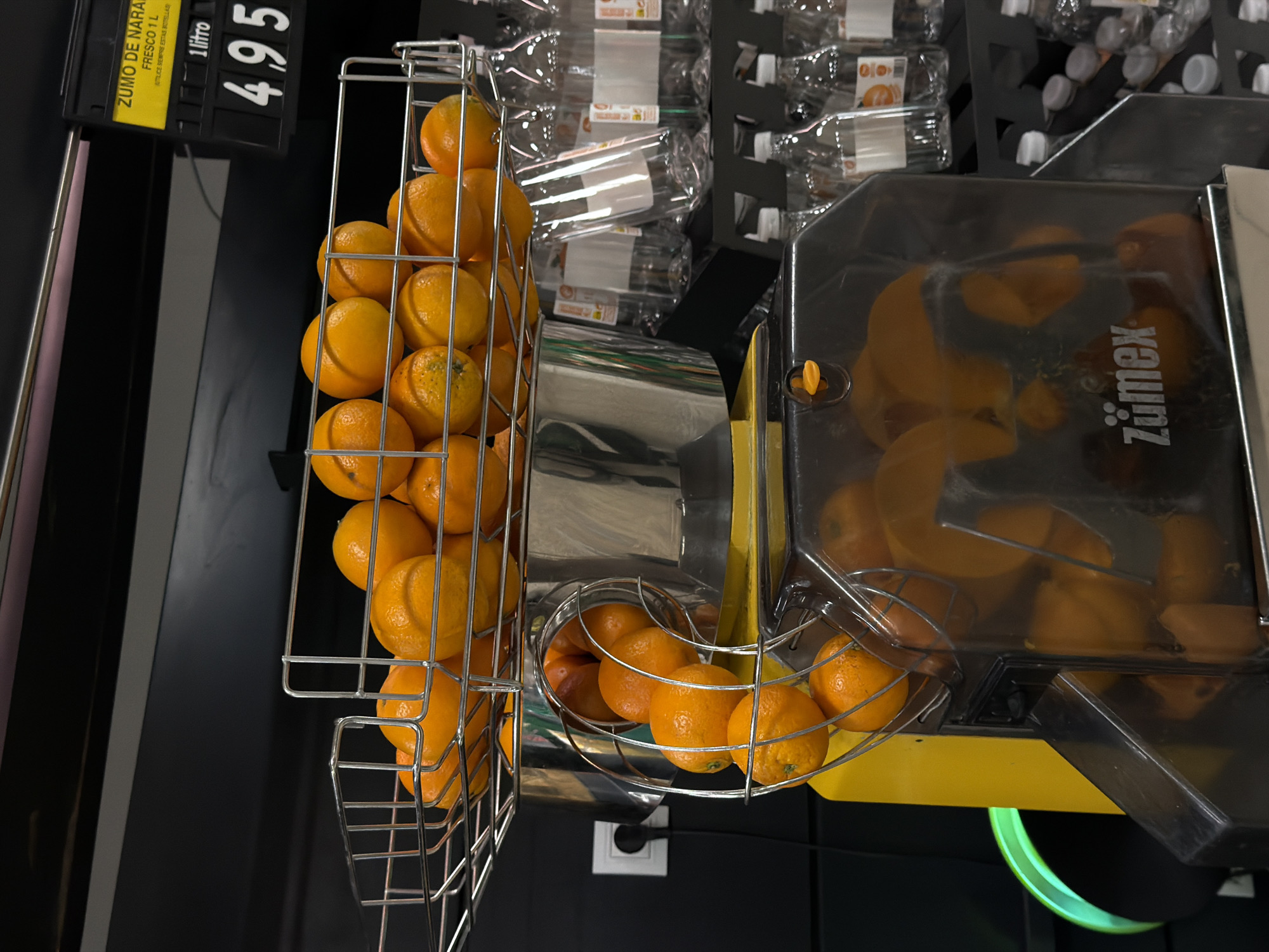 This media file captures a self-service fresh orange juice station, likely within a supermarket or grocery store. The scene is dominated by numerous bright orange oranges, neatly stacked in silver wire racks to the left and integrated into the juicing machine on the right.
At the center, a commercial "Zumex" brand juicer is prominently displayed, featuring a transparent top through which several processed orange halves are visible, indicating recent or ongoing operation. Adjacent to the juicer, a cylindrical metal chute feeds oranges into the machine, and another rack holds more fresh oranges ready for juicing.
Above and behind the juicer, a display of empty, clear plastic bottles with white caps is stacked, intended for customers to fill with the freshly squeezed juice.
A price tag, oriented vertically on the left side of the frame, details the product and cost. The text reads: "ZUMO DE NARANJA FRESCO 1 L" (Fresh Orange Juice 1 L), "REUTILICE SIEMPRE ESTAS BOTELLAS" (Always reuse these bottles), and clearly displays "4.95" for "1 litro" (1 liter), strongly suggesting a price of 4.95 Euros per liter, consistent with the location context of Valencia, Spain. Small text "010" is also visible on some of the bottle labels or the price tag.
The background is predominantly dark, providing a stark contrast to the vibrant oranges and the metallic elements of the juicer and racks. The overall impression is one of freshness and convenience for customers seeking freshly made orange juice.