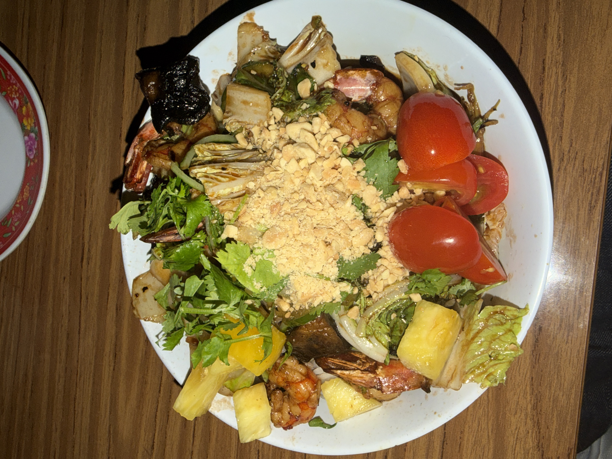A white bowl containing a shrimp salad is positioned on a wooden table. The salad includes shrimp, tomatoes, leafy greens (possibly cilantro), pineapple chunks, and chopped peanuts. Another plate with a red rim is adjacent to the bowl. The scene is indoors and is potentially located in a restaurant in Buenos Aires, Argentina.