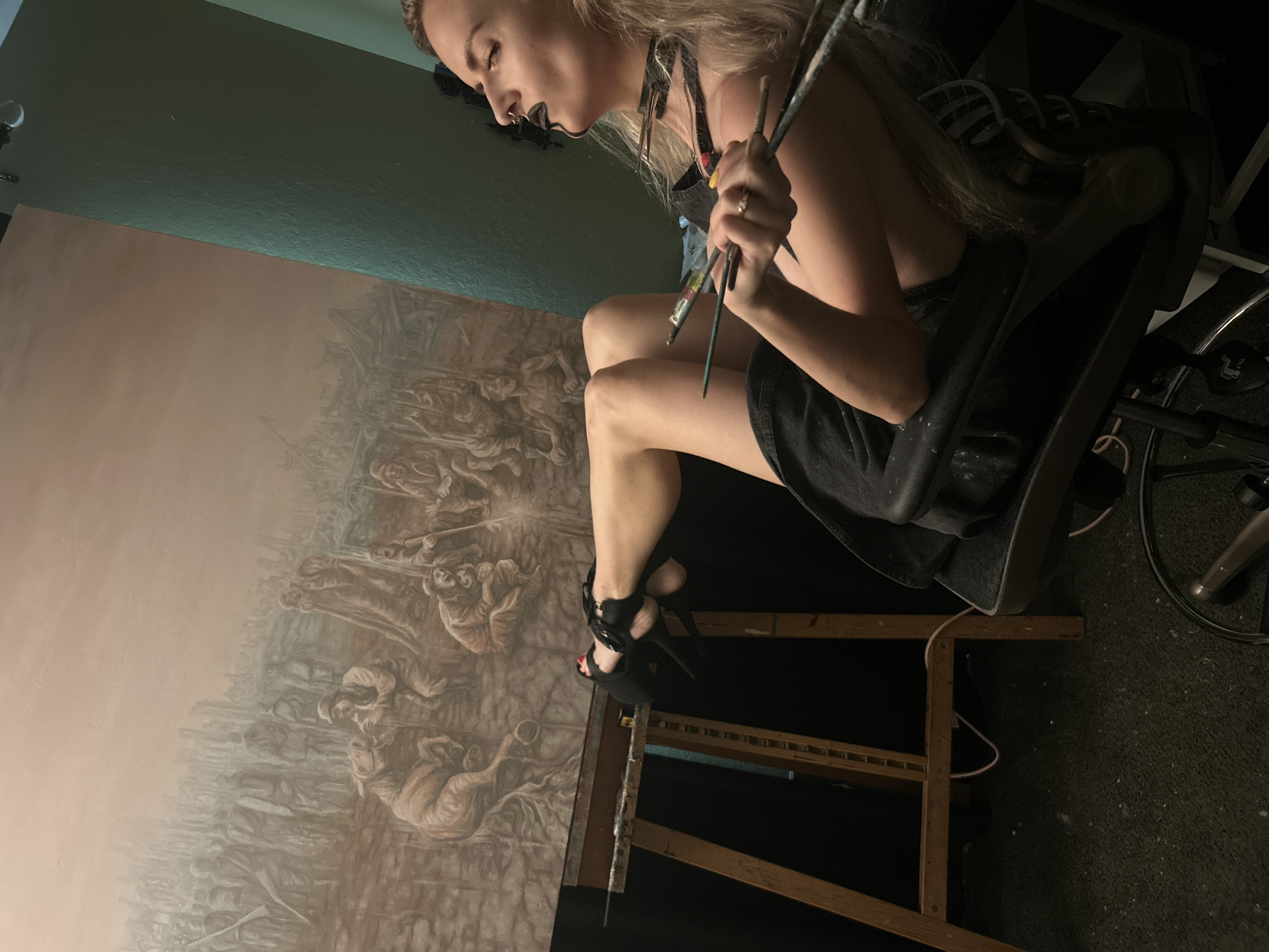 A blonde woman with black lipstick and a black choker is seated in a black chair, holding several paintbrushes. She is wearing black shorts and black high-heeled sandals. Her legs are crossed, and her left foot is resting on the edge of a wooden easel. A large, detailed painting depicting a chaotic scene with many figures and possibly horses is displayed on the easel. The painting has a muted, earthy color palette. The room appears to be a studio, with dark walls and a carpeted floor. The lighting is dim, with a spotlight illuminating the painting and the artist. The overall impression is of an artist in the midst of her creative process. This scene is located in Cape Coral, United States of America.